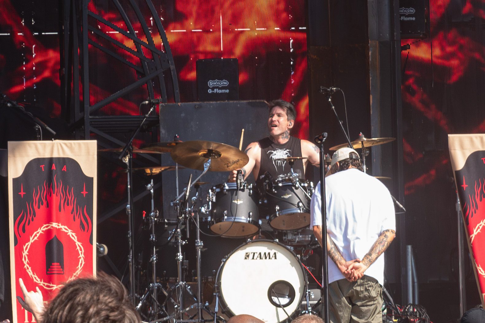 A drummer energetically playing on stage with a band at a music festival, featuring a backdrop of vibrant graphics and a banner with a bold design.