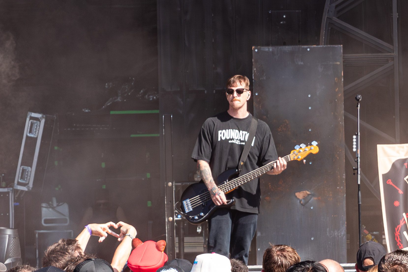 A bassist performing on stage at a music festival, wearing sunglasses and a black t-shirt, with a crowd in the foreground.