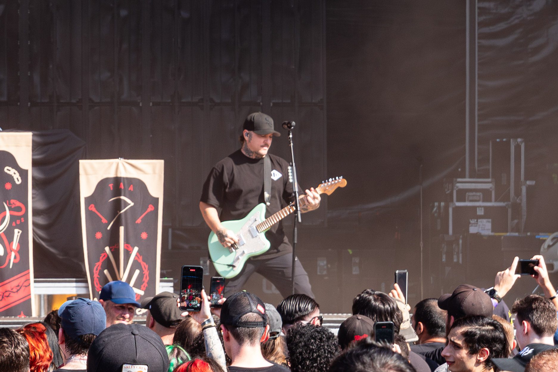 A guitarist on stage performing with a mint green guitar, in front of an audience, as banners with designs are displayed behind him.