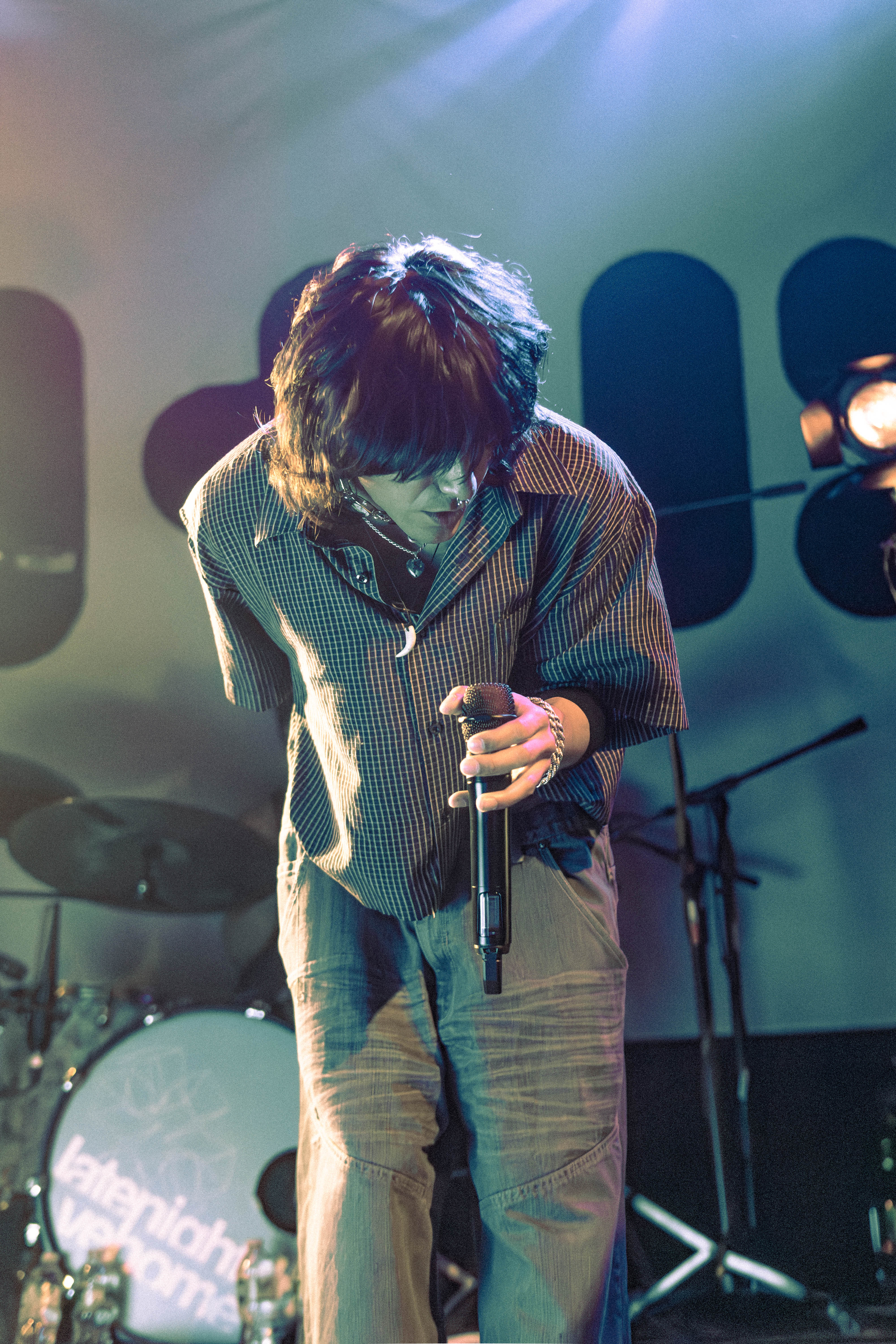 A member of the band late night drive home performing on stage, holding a microphone and wearing a casual plaid shirt. The stage is illuminated with colorful lights.