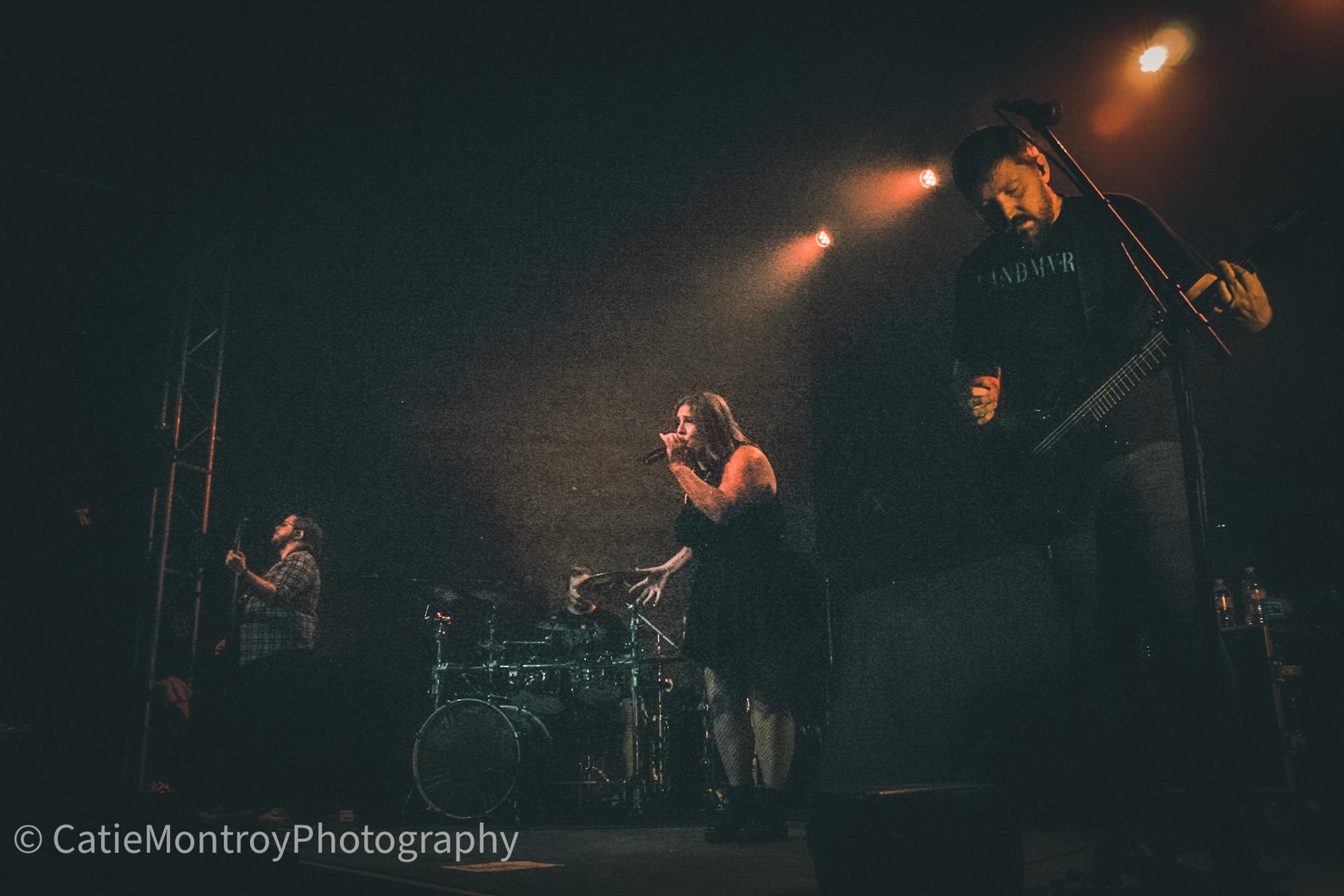 A live performance of the band Wabash & Lake at a concert, featuring a female lead singer on stage with a microphone, accompanied by a guitarist and a drummer in the background with stage lights illuminating the scene.
