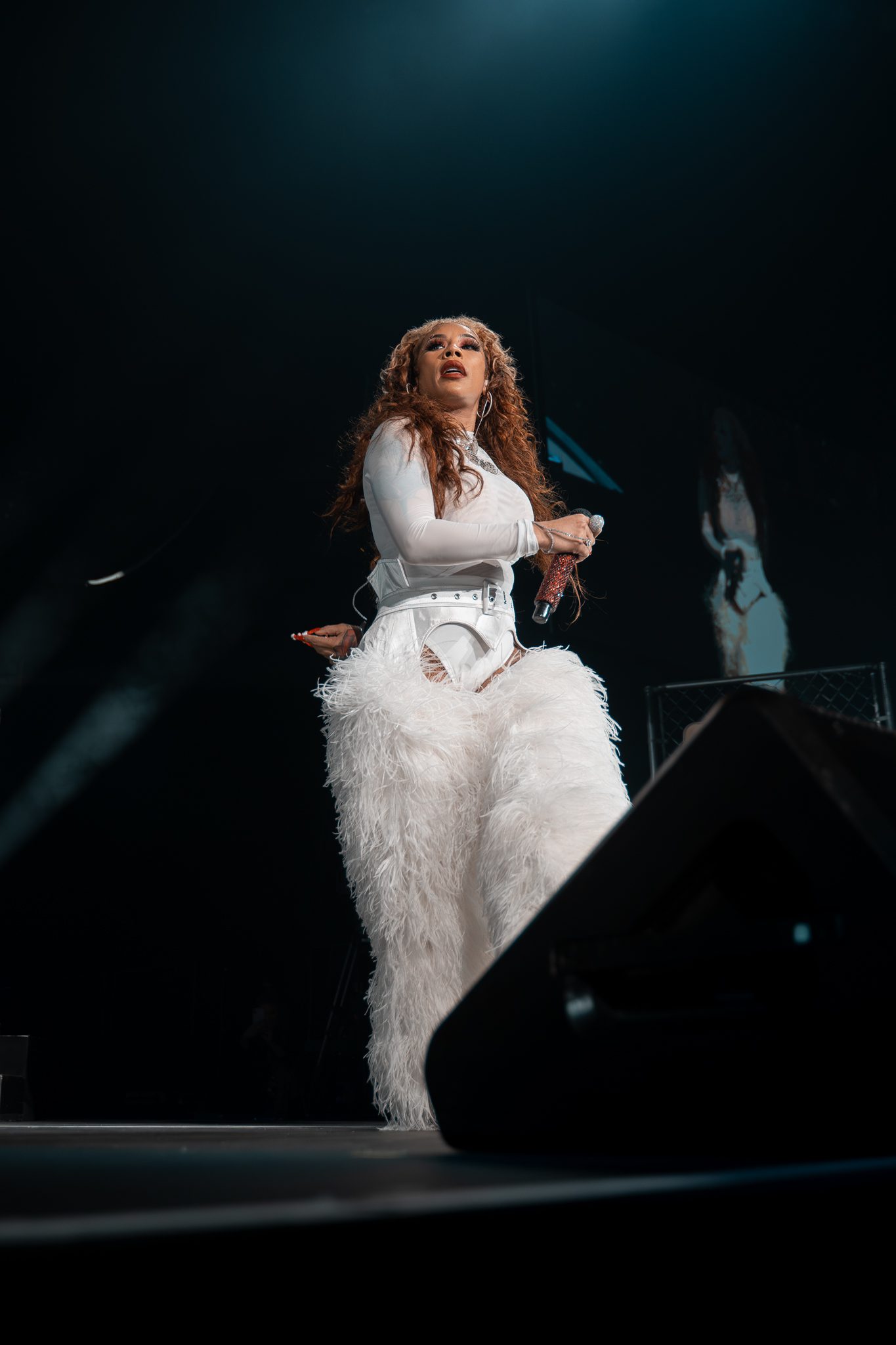 Keyshia Cole performing on stage, wearing a full white outfit with fluffy, feathery pants, holding a microphone and engaging with the audience.