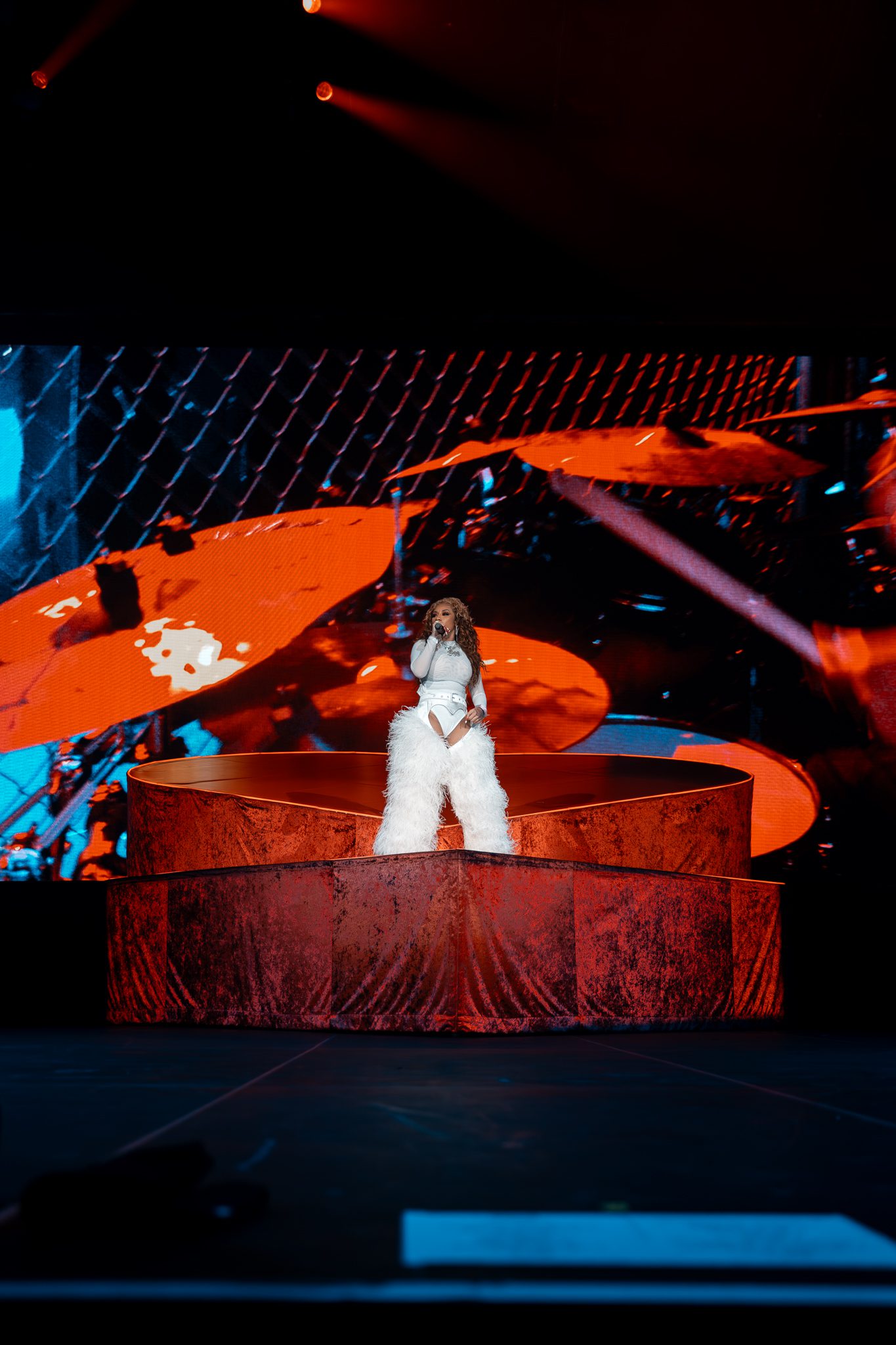 Keyshia Cole performing on stage in a white outfit with fluffy pants, standing on a heart-shaped platform with a colorful backdrop.