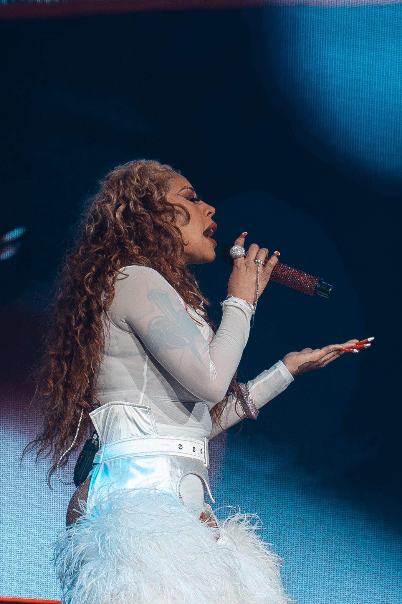 Keyshia Cole performing on stage, wearing a white outfit with fluffy pants, holding a microphone while singing.