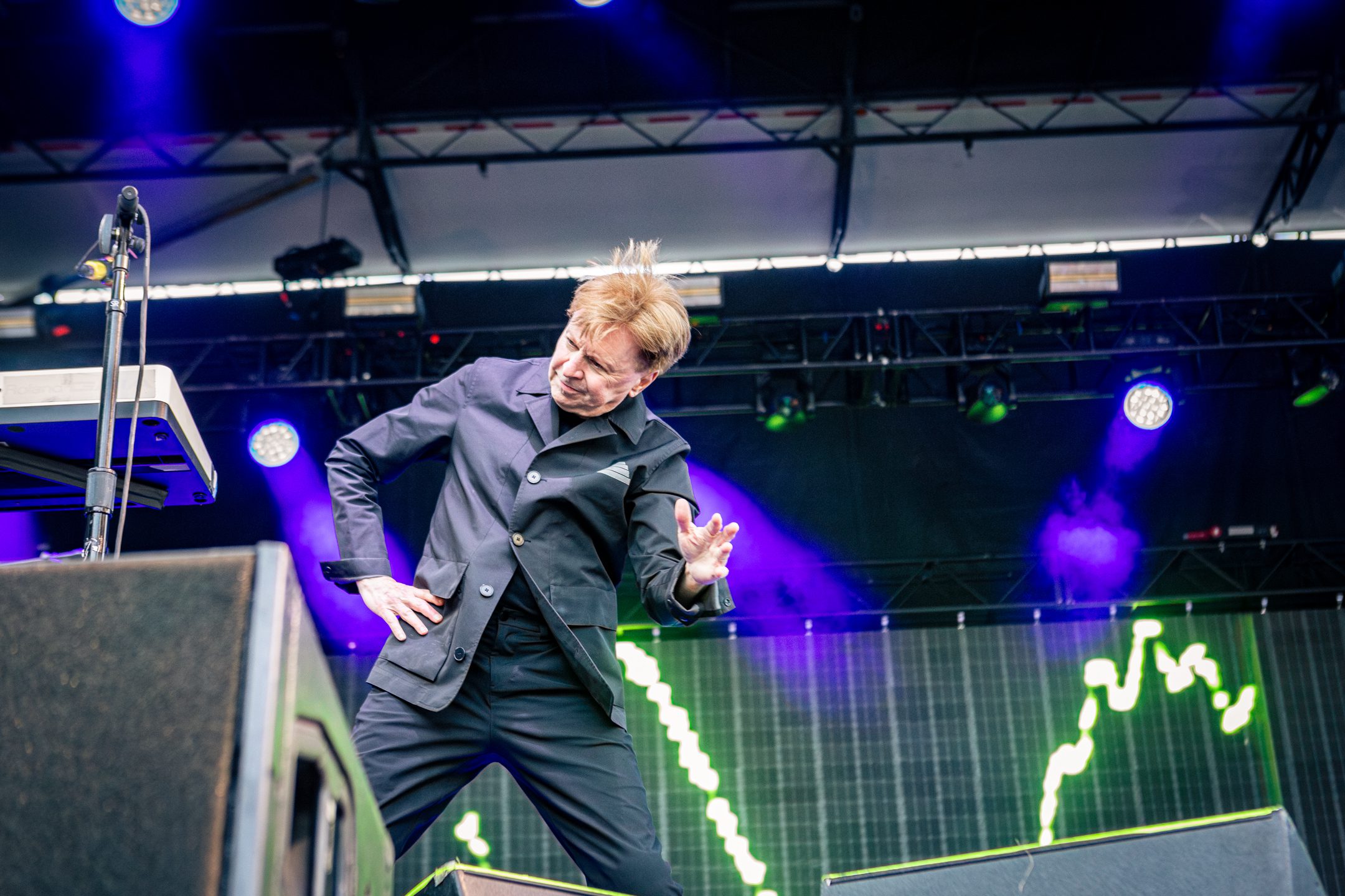 A performer on stage passionately engaging with the audience, wearing a black jacket against a colorful backdrop of lighting.