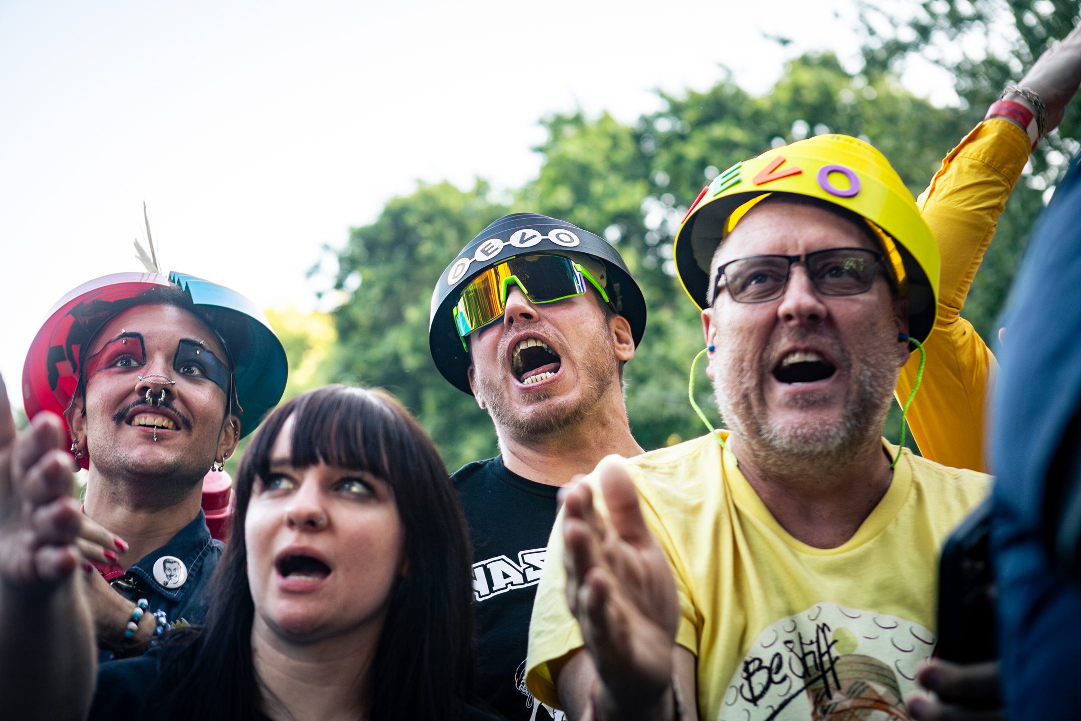 A group of enthusiastic festival-goers wearing colorful helmets and expressing excitement, with one person wearing makeup and facial piercings, enjoying the performance.