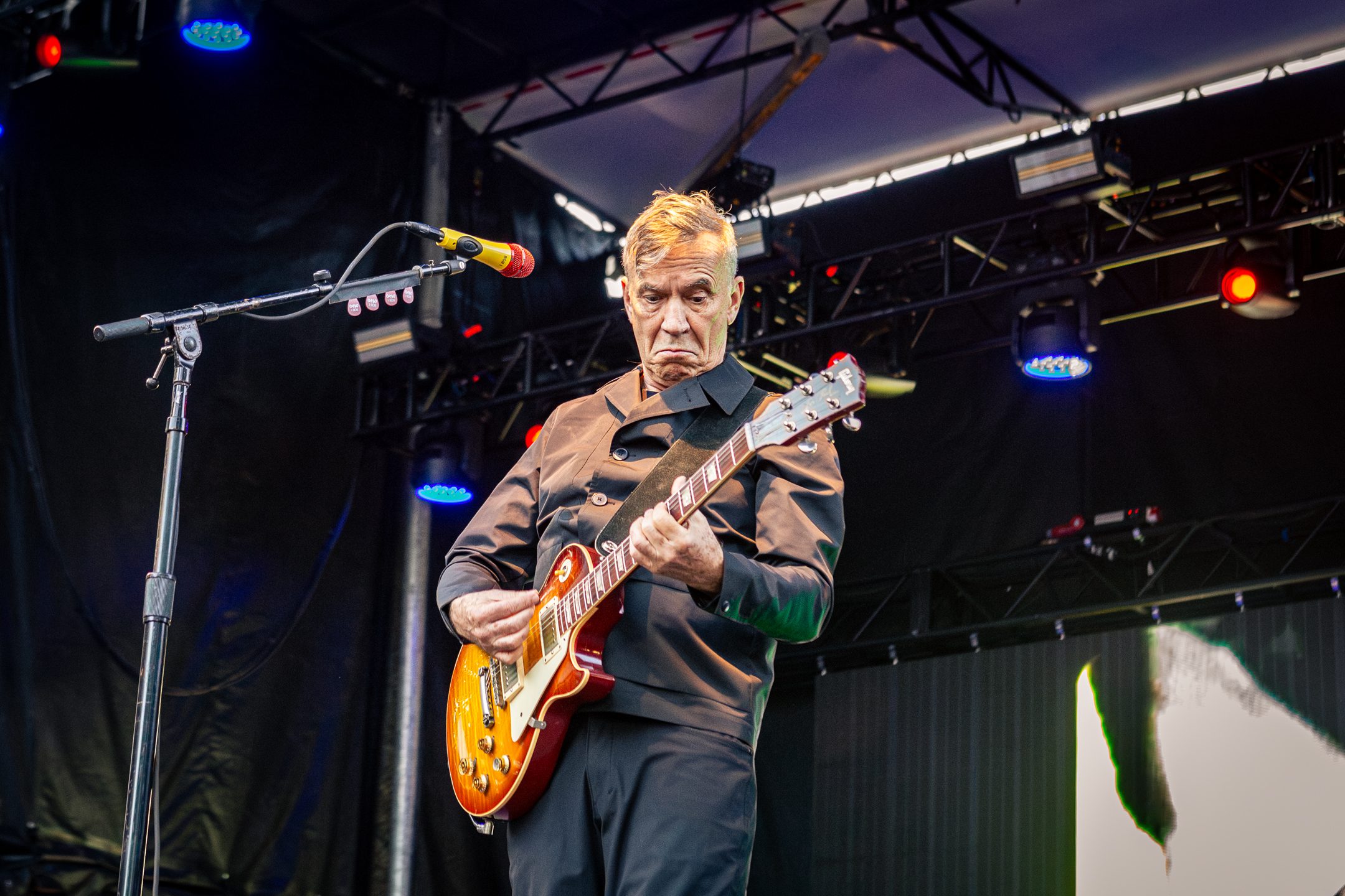 A musician performing on stage, wearing a black coat and playing an electric guitar, with intense expression and colorful stage lighting in the background.