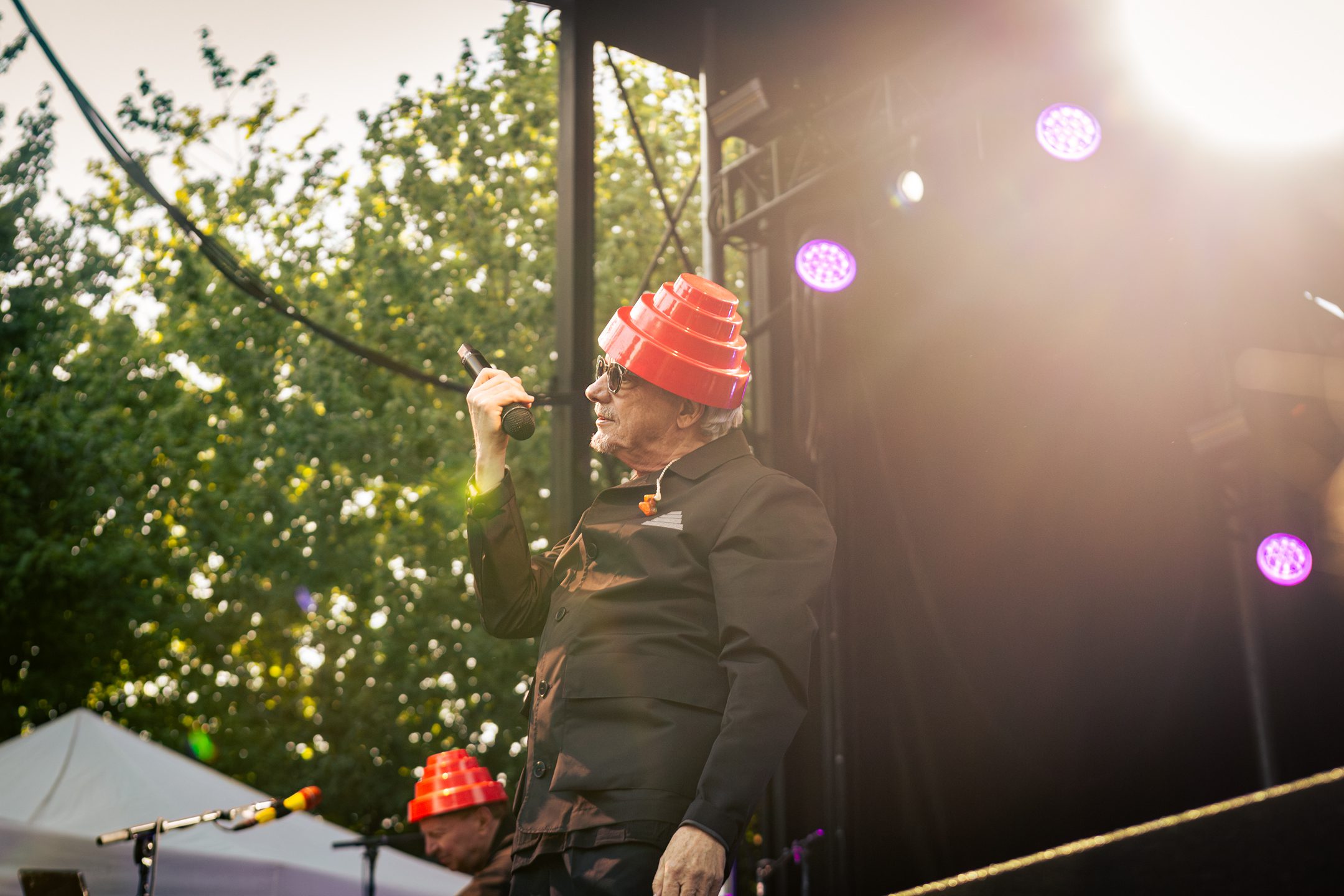 A performer wearing a red energy dome hat and black outfit stands on stage, holding a microphone, with trees and stage lights in the background.