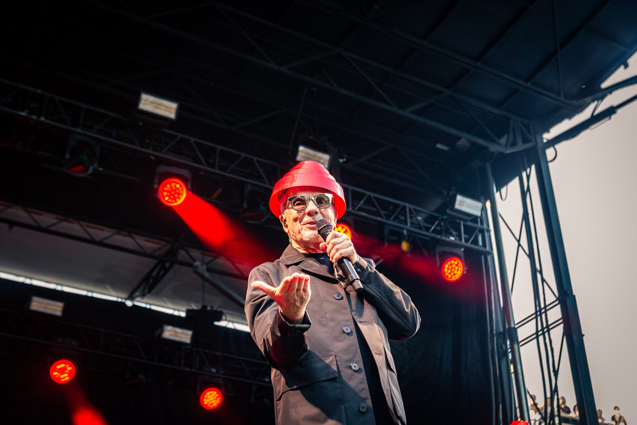 A performer in a black jacket and red hat engages the audience on stage during a music festival, with red stage lights illuminating the scene.