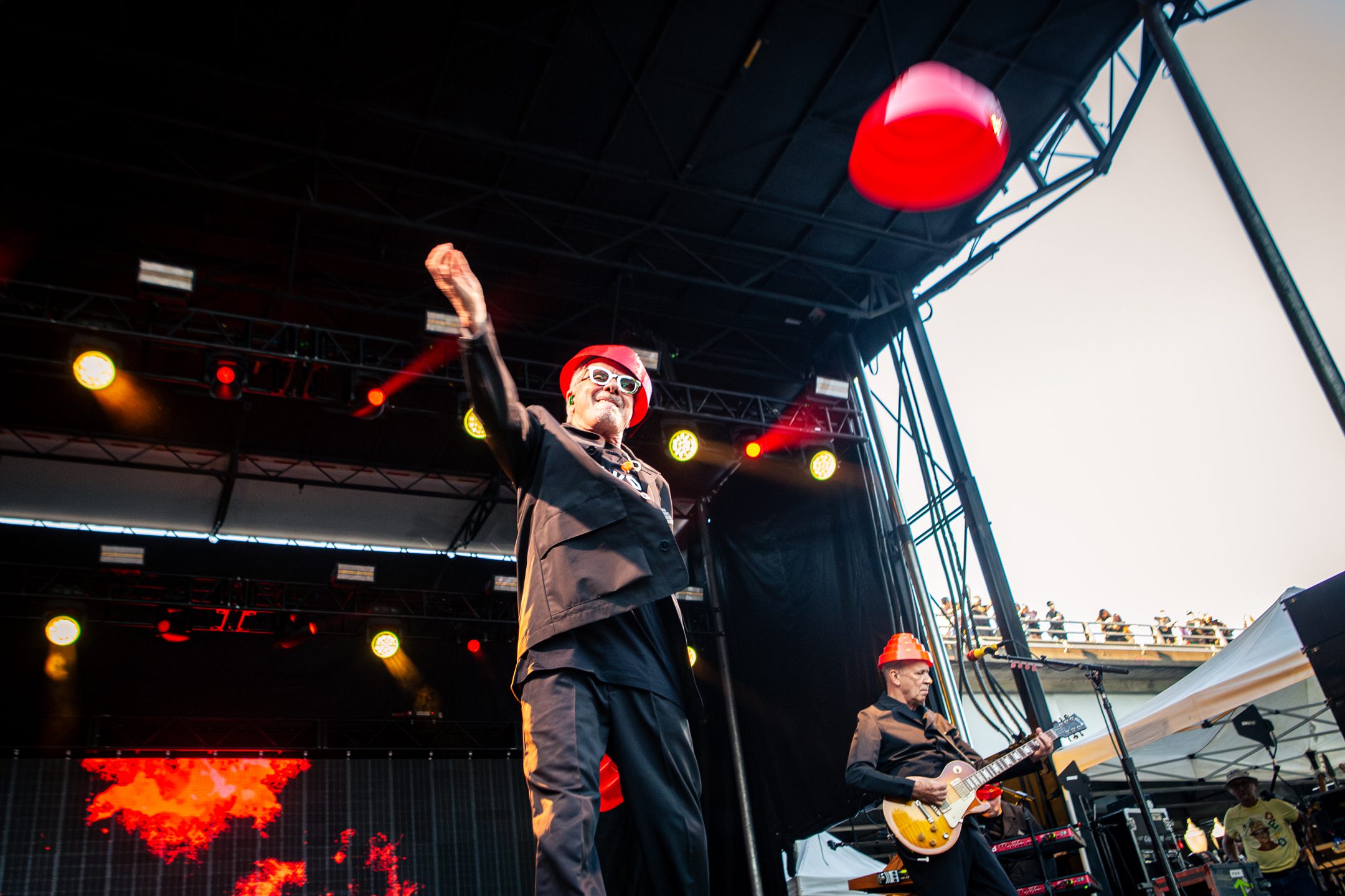 A performer wearing a red hat and sunglasses throws a red object during a live music performance on stage, while a guitarist plays beside him.