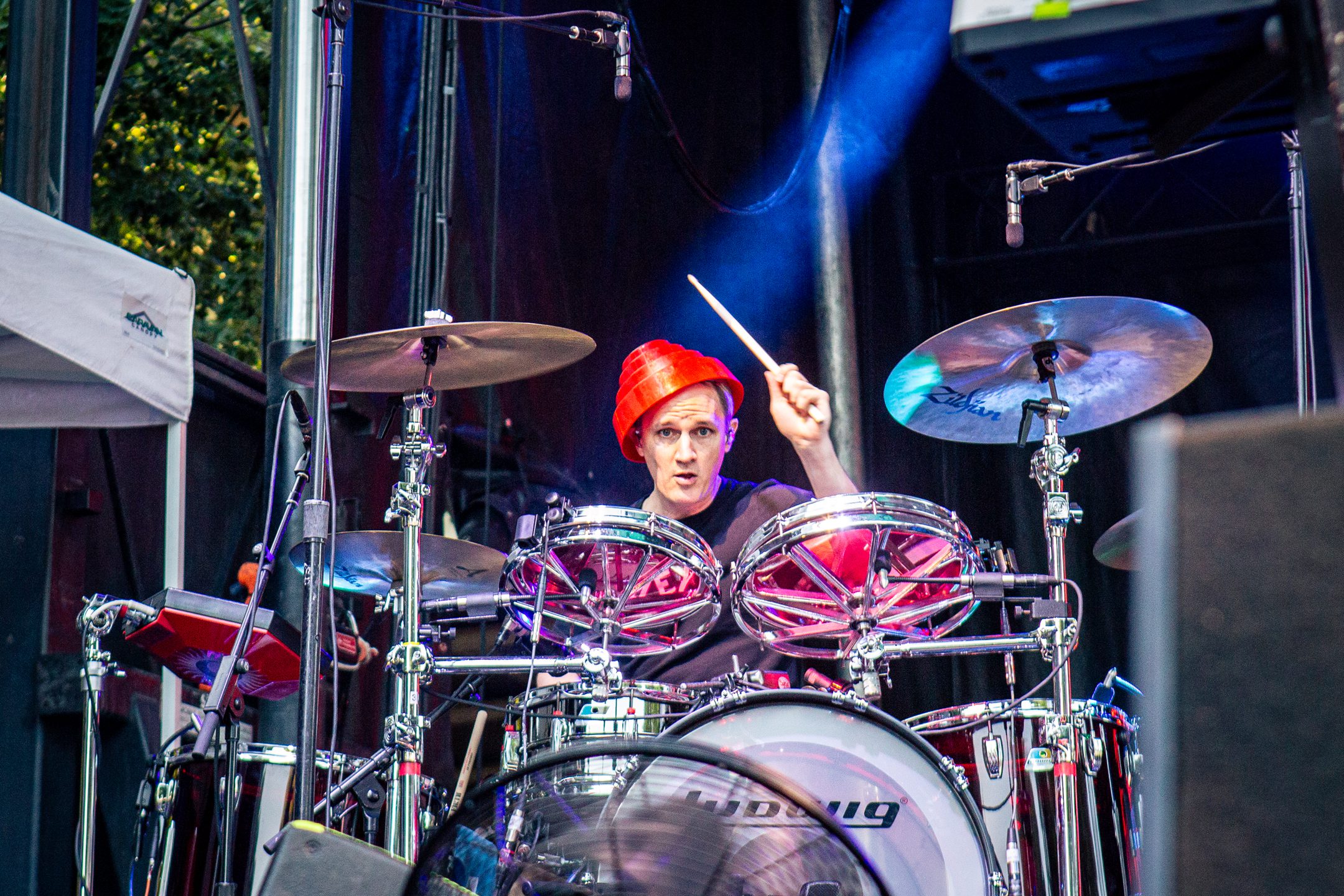 Drummer playing energetic set on stage, wearing a red hat, amidst drum kit and vibrant lighting.