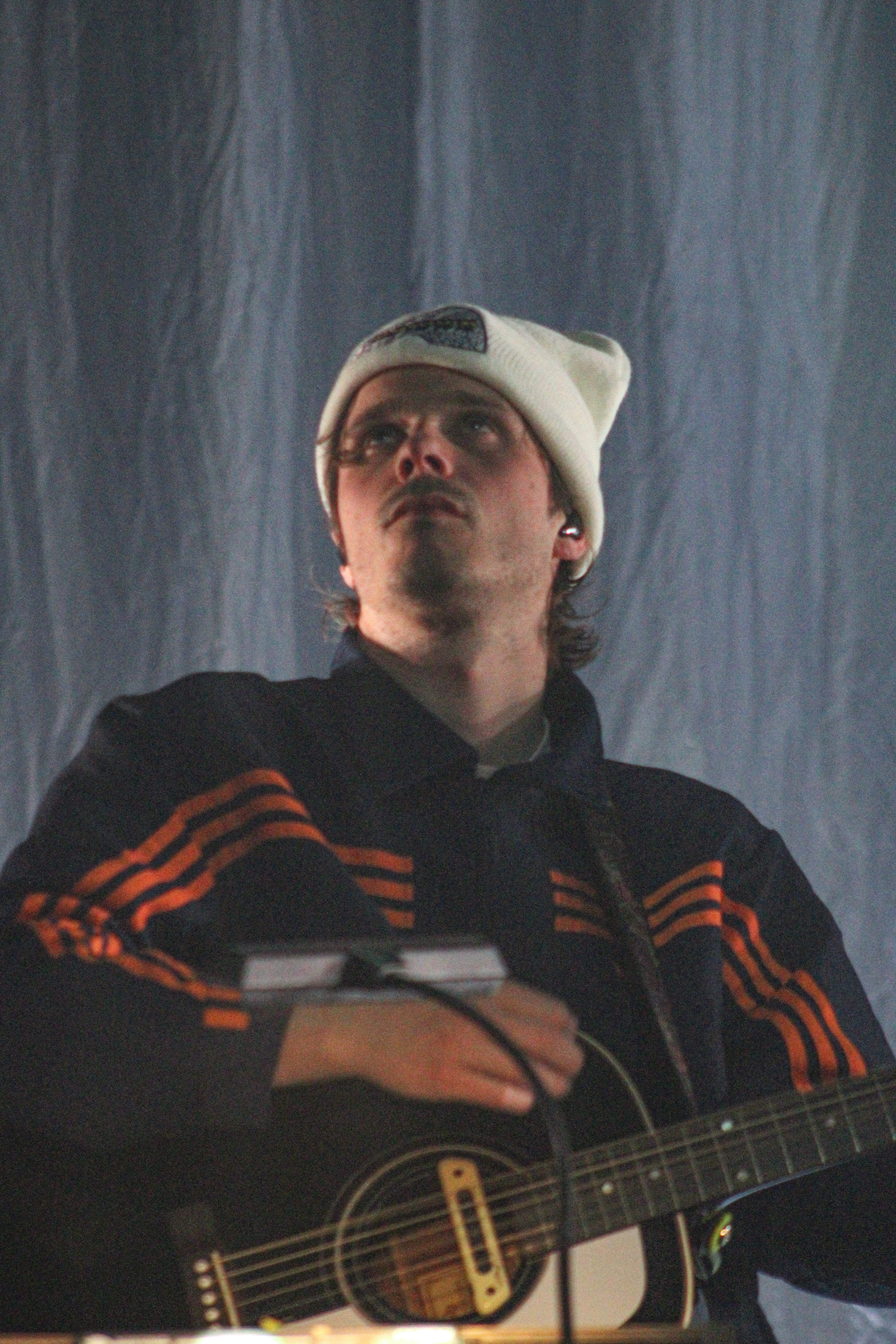 A musician wearing a beanie and a jacket with orange stripes, playing an acoustic guitar on stage, with a focused expression.