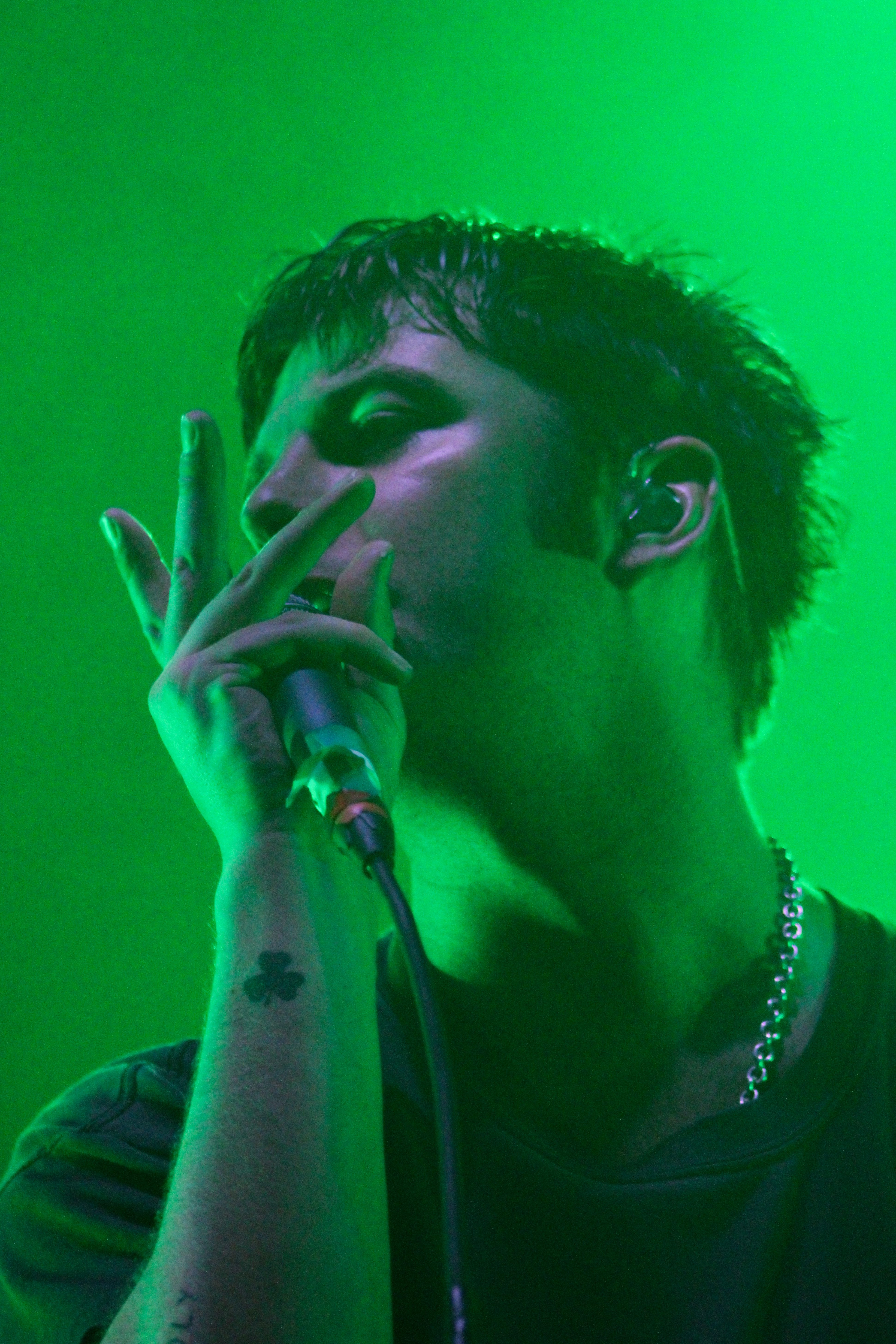Close-up of a male vocalist performing on stage under green lighting, holding a microphone and expressing emotion through gestures.