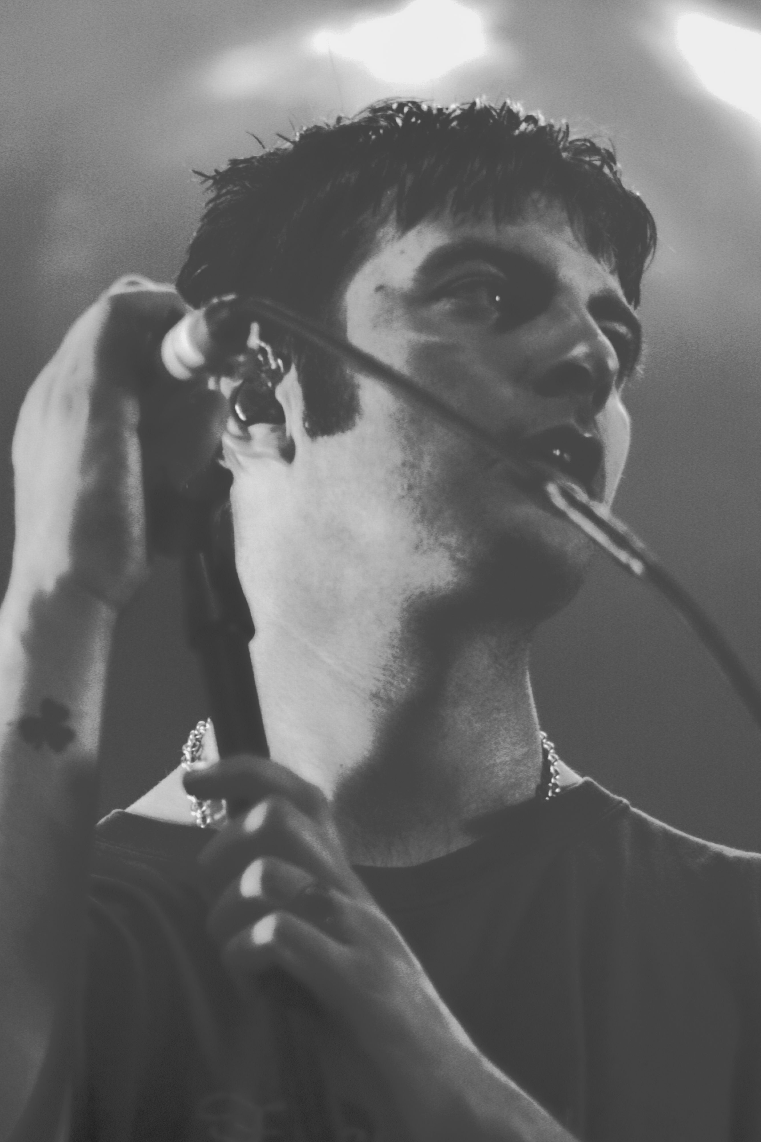 A black and white close-up of a male singer holding a microphone, passionately performing on stage. He has short hair and a small tattoo visible on his arm.