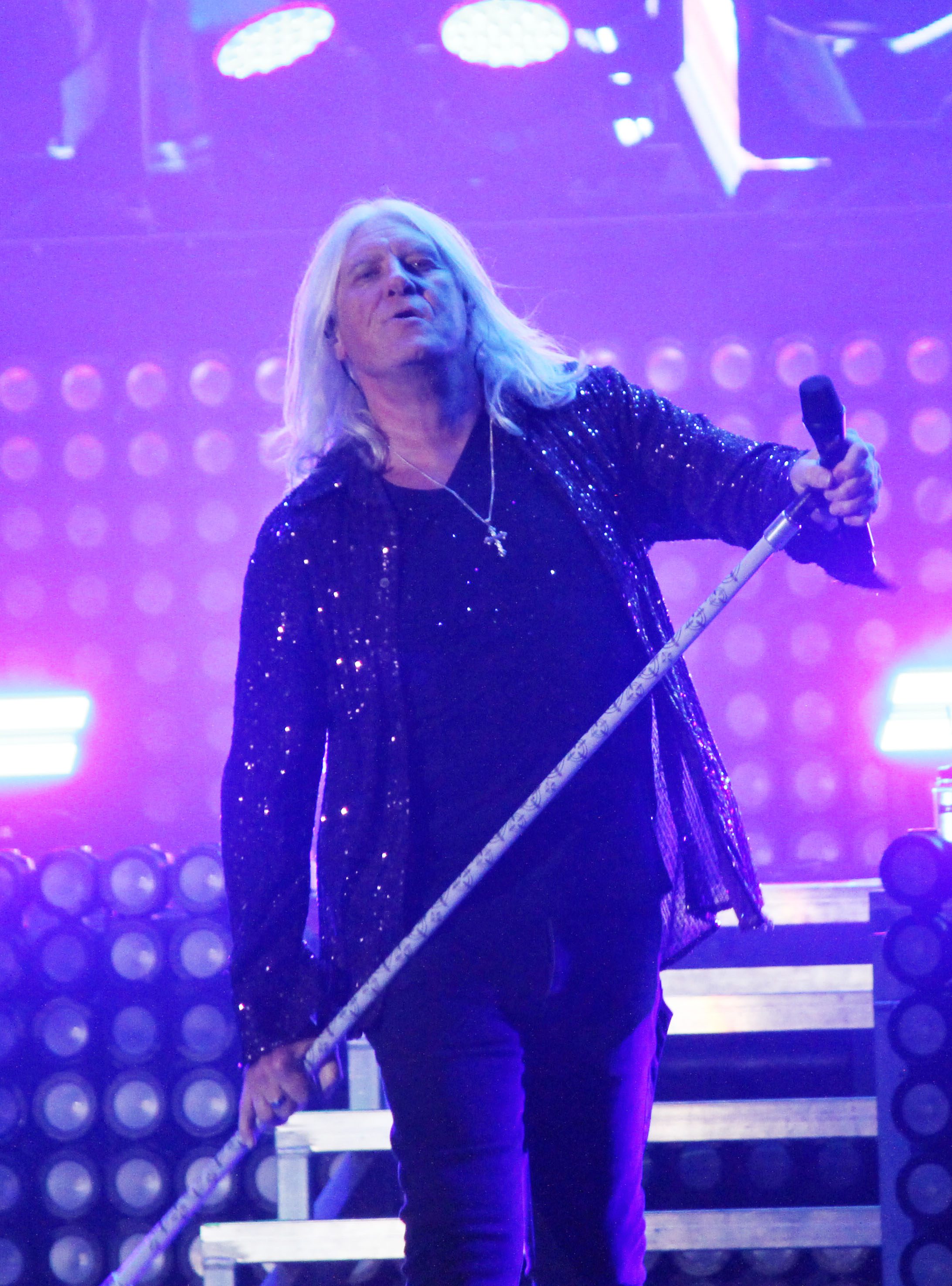 Lead singer Joe Elliott of Def Leppard performing on stage with a microphone, dressed in a sparkly jacket under purple stage lights.