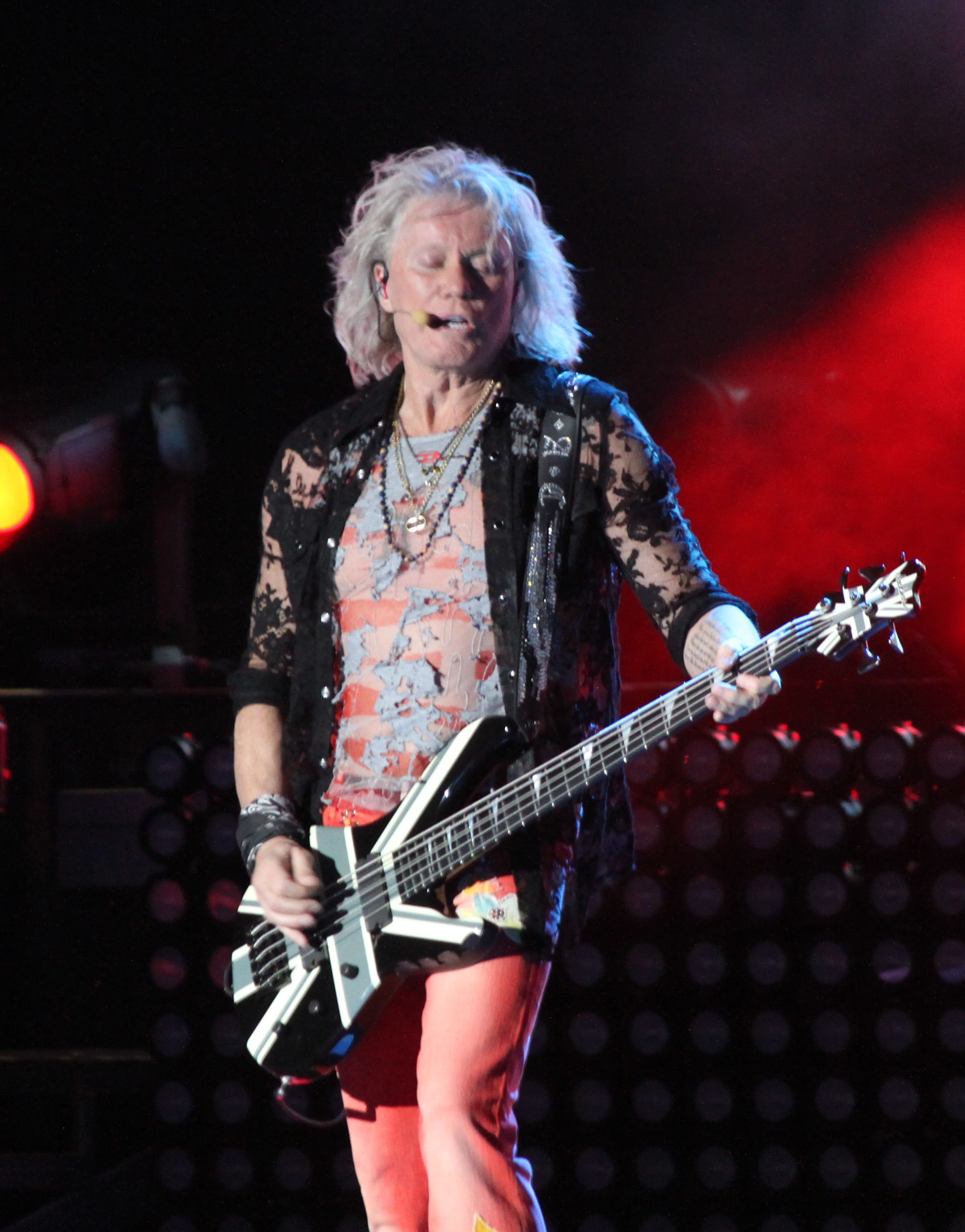 Musician performing on stage with a distinct bass guitar, wearing a stylish outfit and surrounded by concert lights.