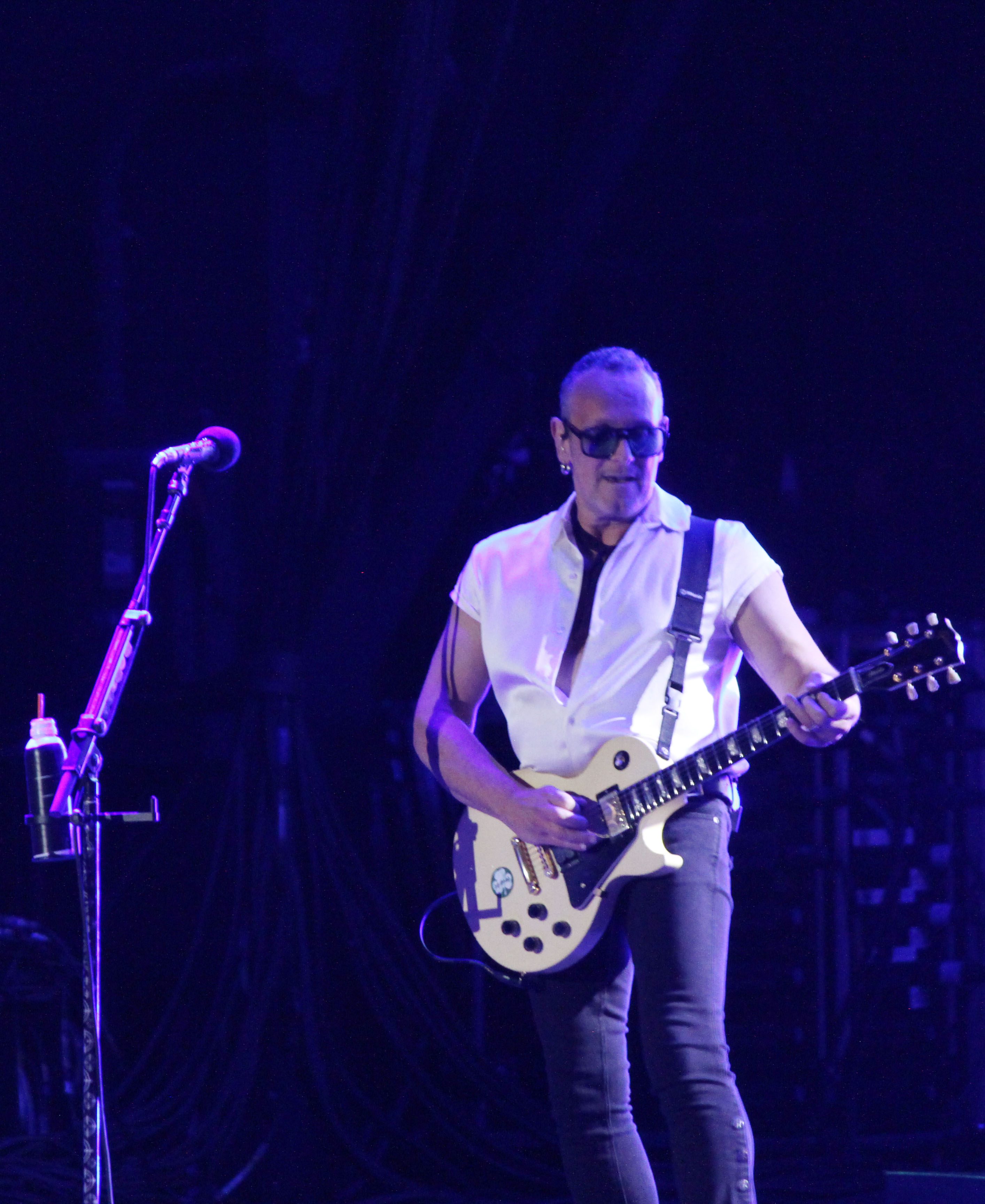 Guitarist performing on stage at a concert with a white guitar and sunglasses, wearing a short-sleeved, button-up shirt.