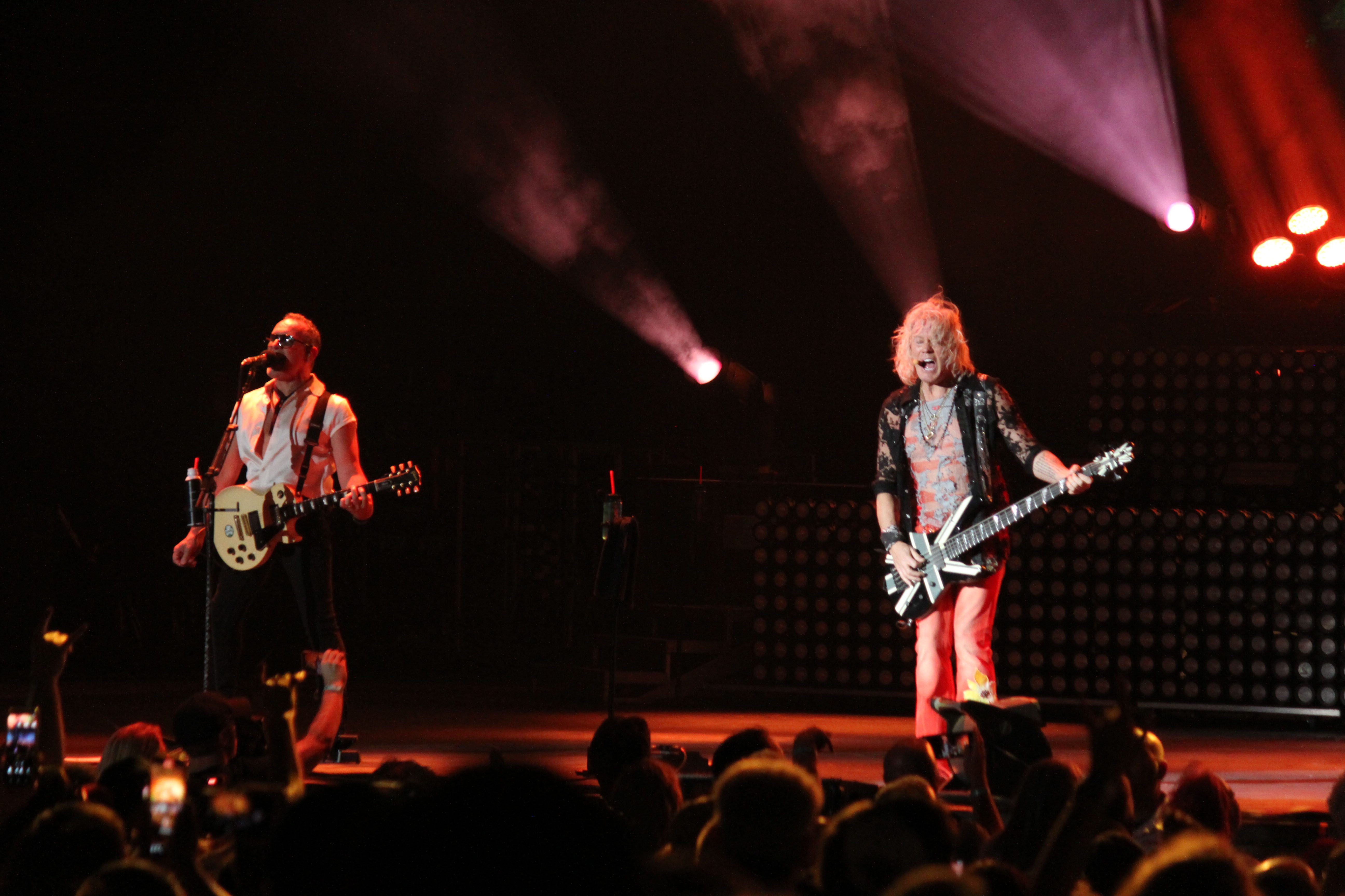 Def Leppard performing on stage with one guitarist playing an electric guitar and another member singing, surrounded by vibrant stage lights and an enthusiastic crowd.
