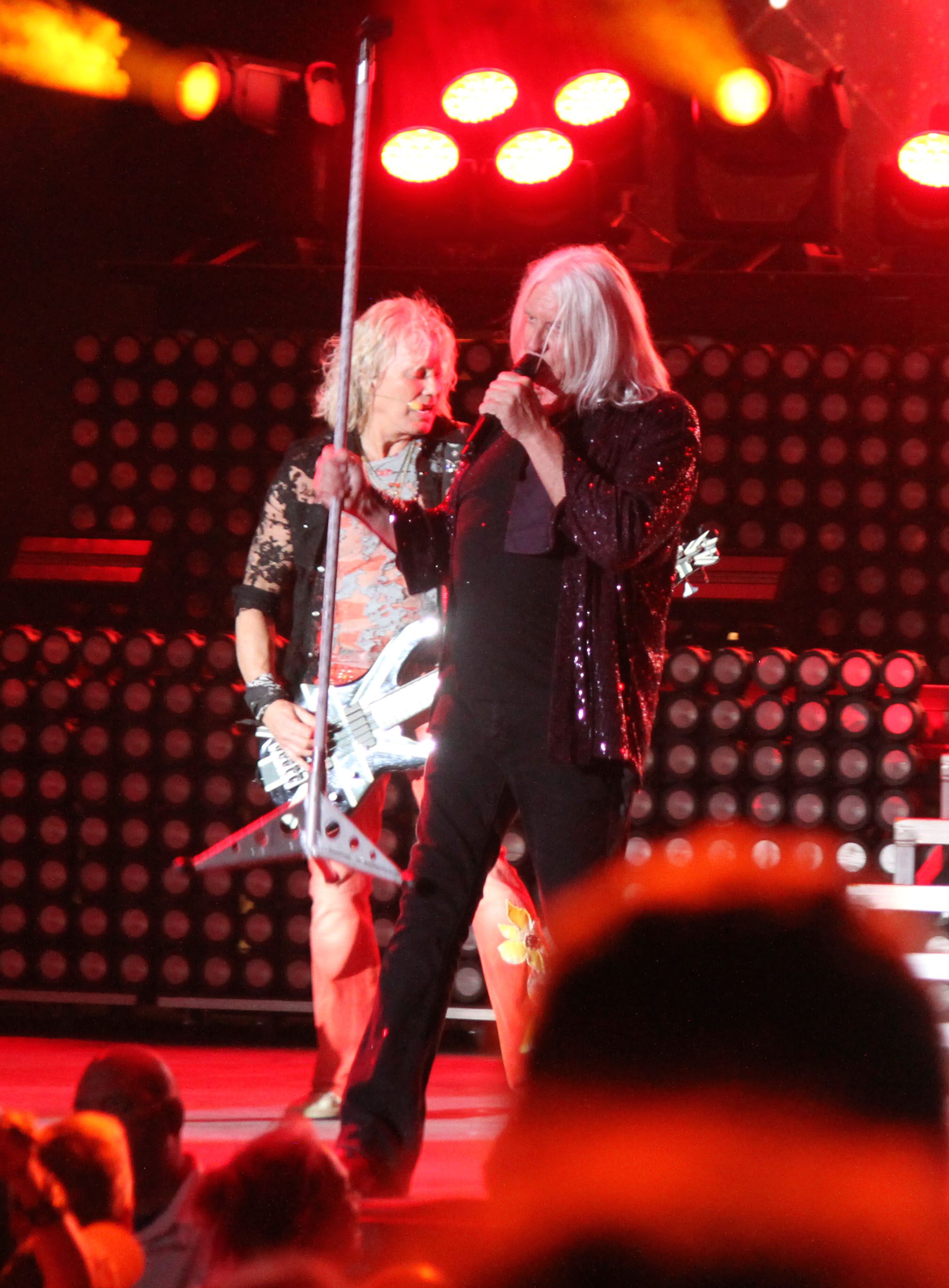 Def Leppard performing live on stage, with the lead singer in a sparkly jacket and a guitarist in a lace shirt, under vibrant stage lights.