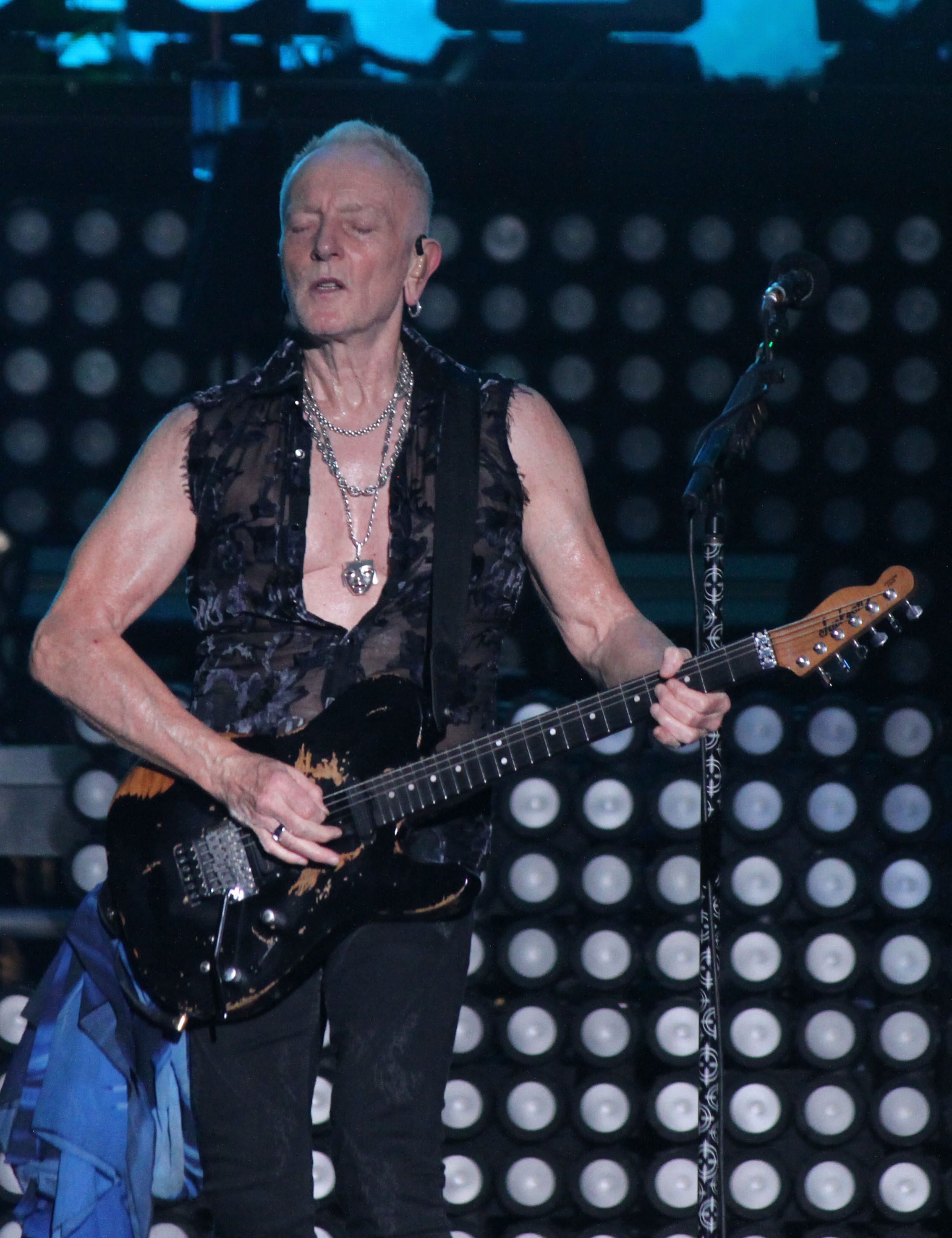 Musician performing on stage, wearing a sleeveless top and holding a guitar, with eyes closed while playing.