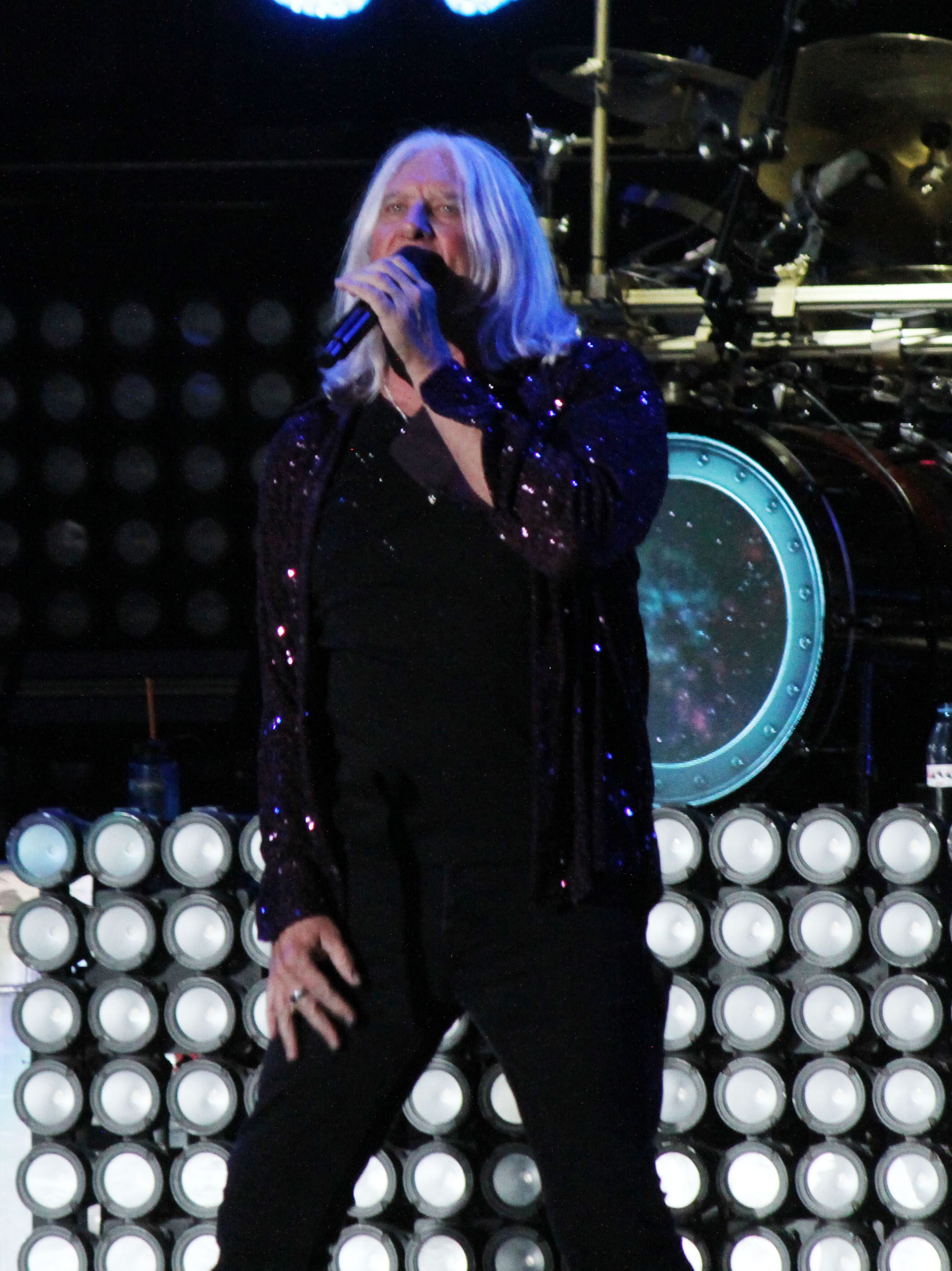 A lead singer performing on stage with long hair and a sparkly jacket, microphone in hand, surrounded by bright stage lights and a backdrop of round lights.