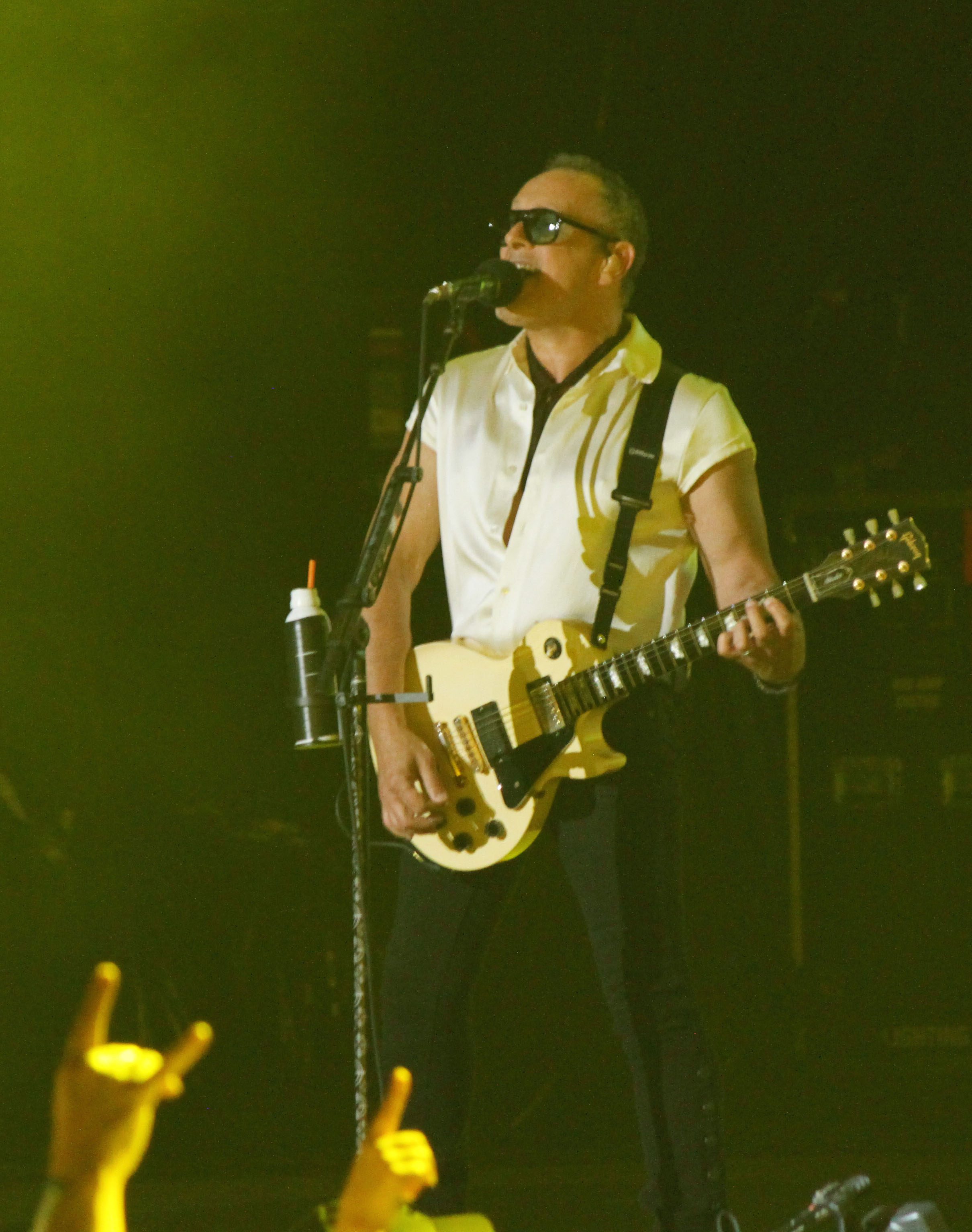 A lead guitarist wearing sunglasses performs on stage with a white guitar, while a crowd shows enthusiastic hand gestures in the foreground.