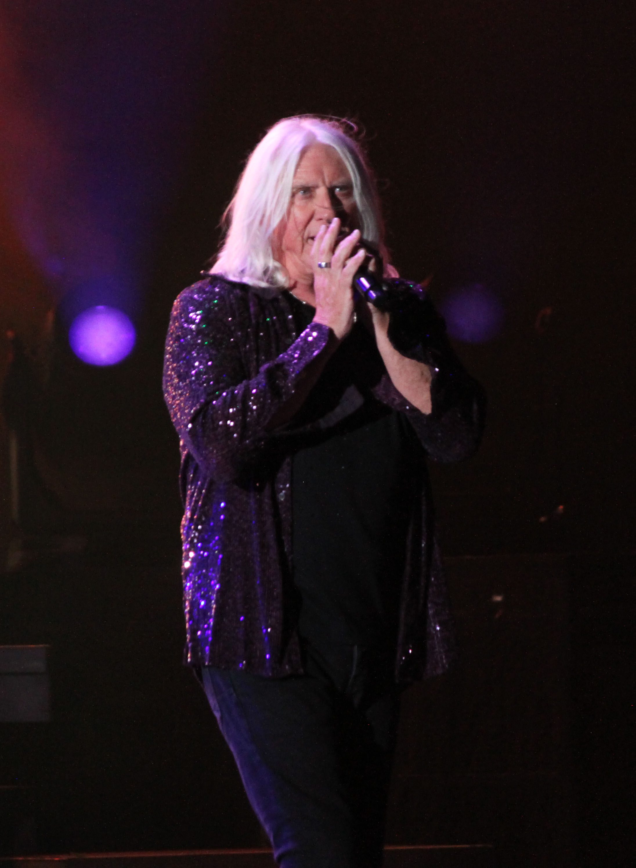 A lead singer performing on stage, wearing a sparkly black jacket under colorful stage lights.