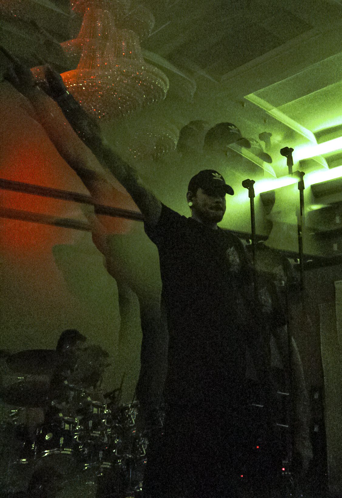 A darkly lit stage with a performer holding his arms up, illuminated by green and orange lights, with a drummer partially visible in the background.