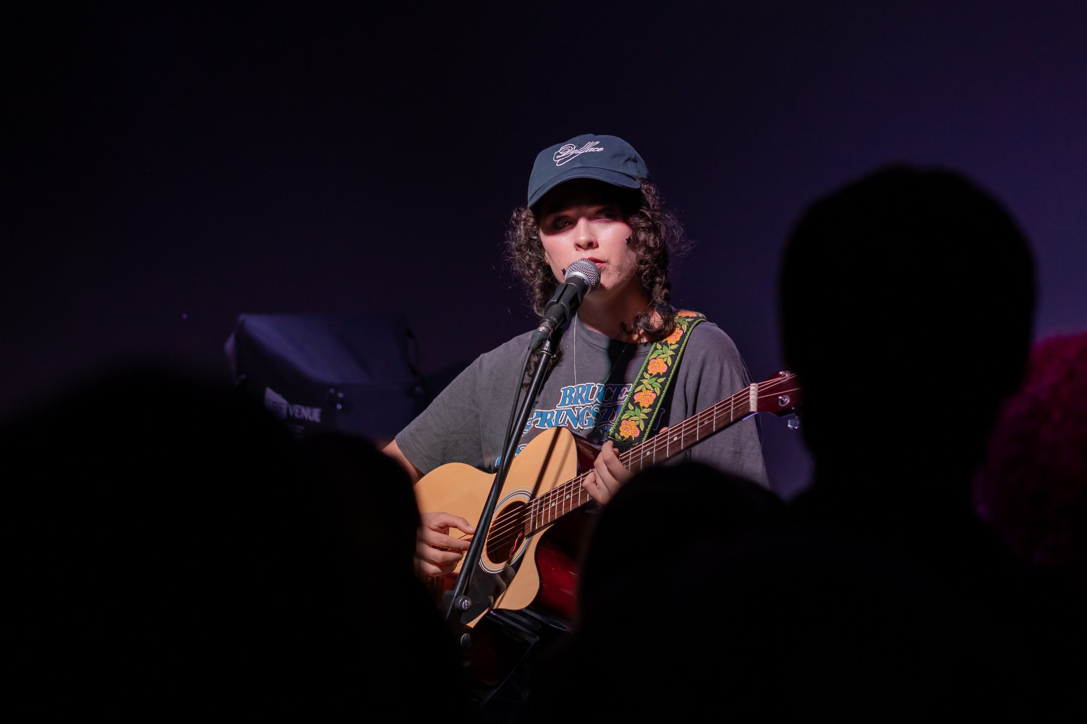 A musician performing on stage, playing an acoustic guitar and singing into a microphone, with an audience silhouette in the foreground.