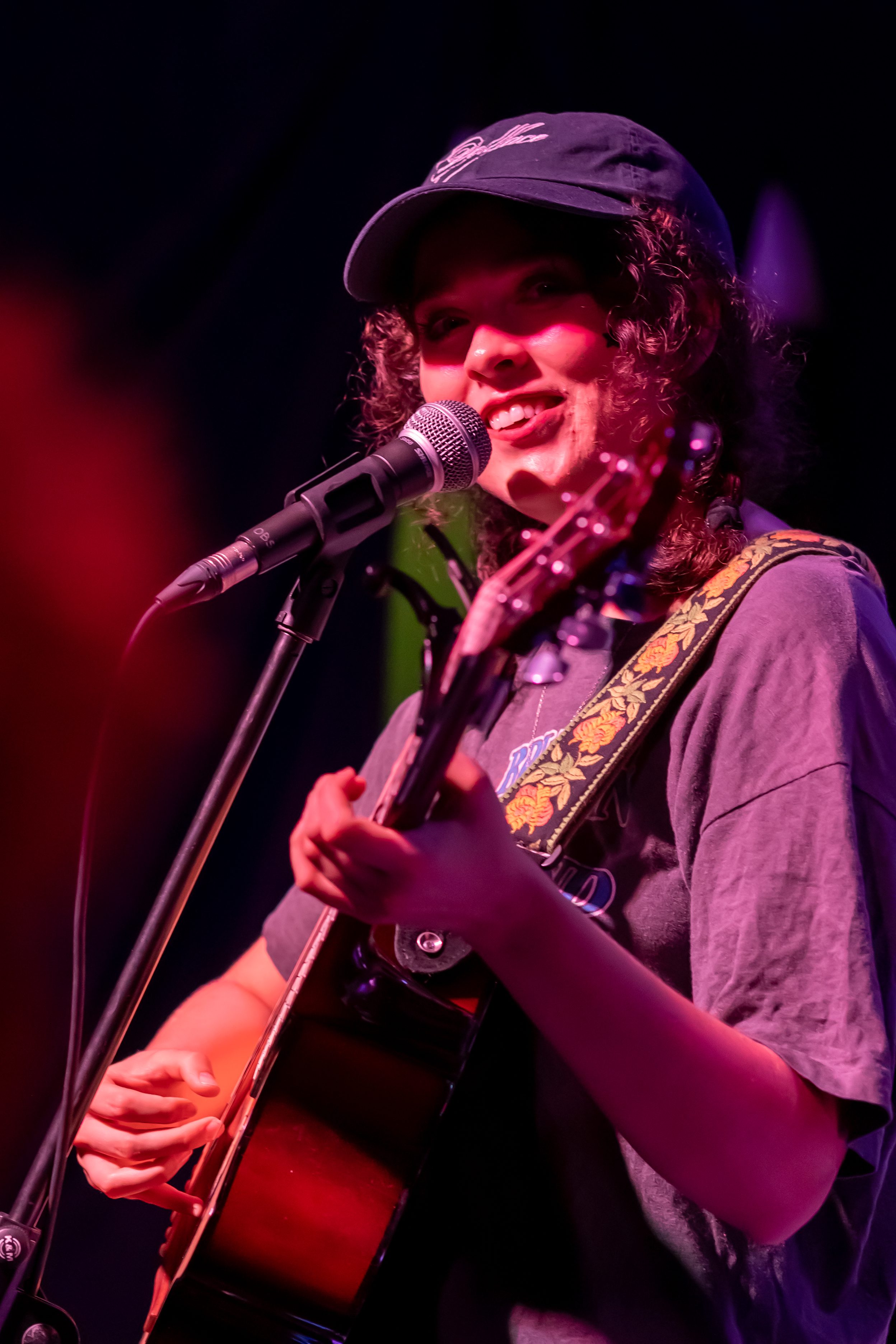 A musician smiling while playing guitar and singing into a microphone on stage, wearing a cap and a casual t-shirt.