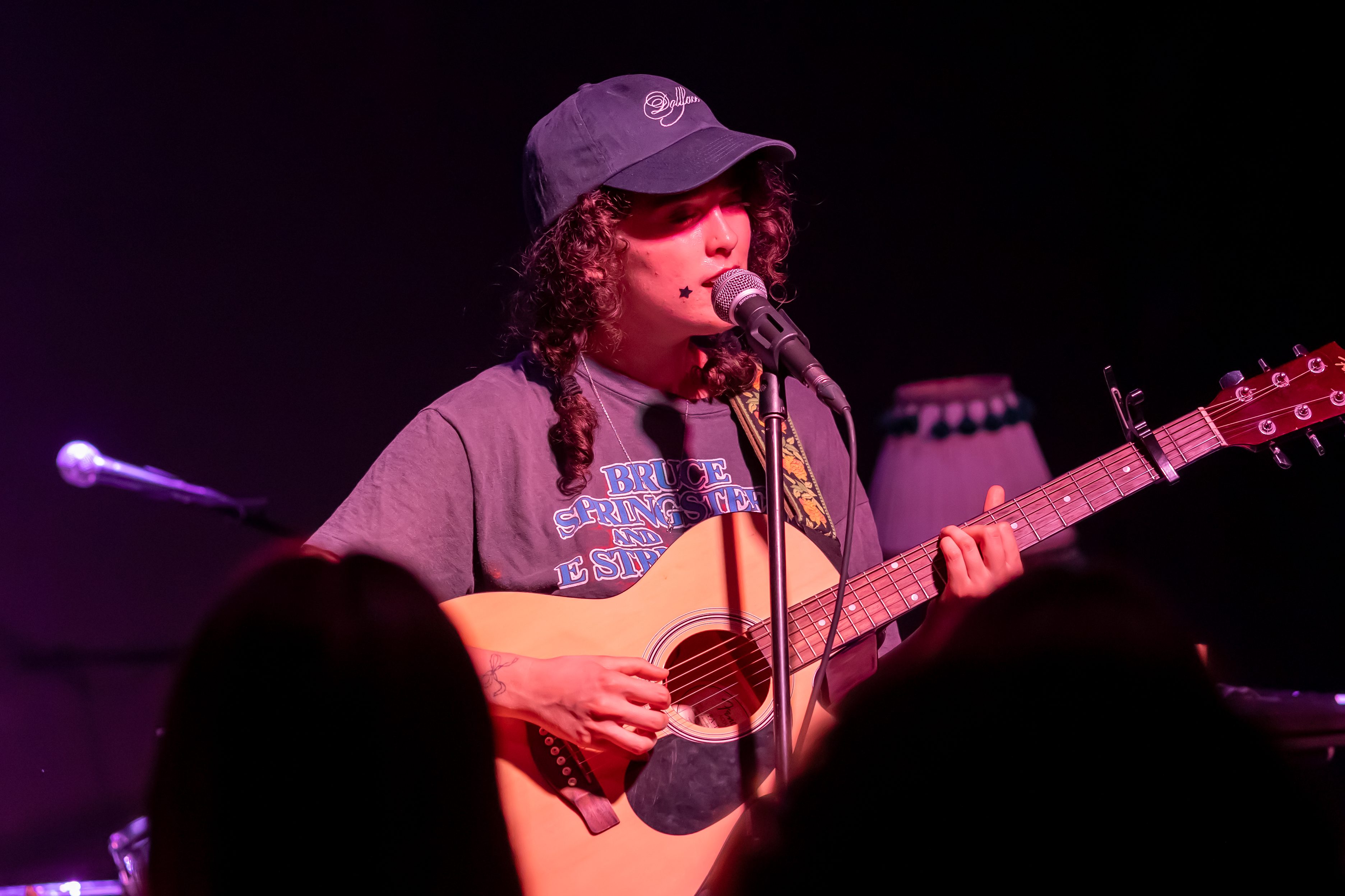 Musician performing on stage, singing into a microphone while playing an acoustic guitar, wearing a cap and a t-shirt.