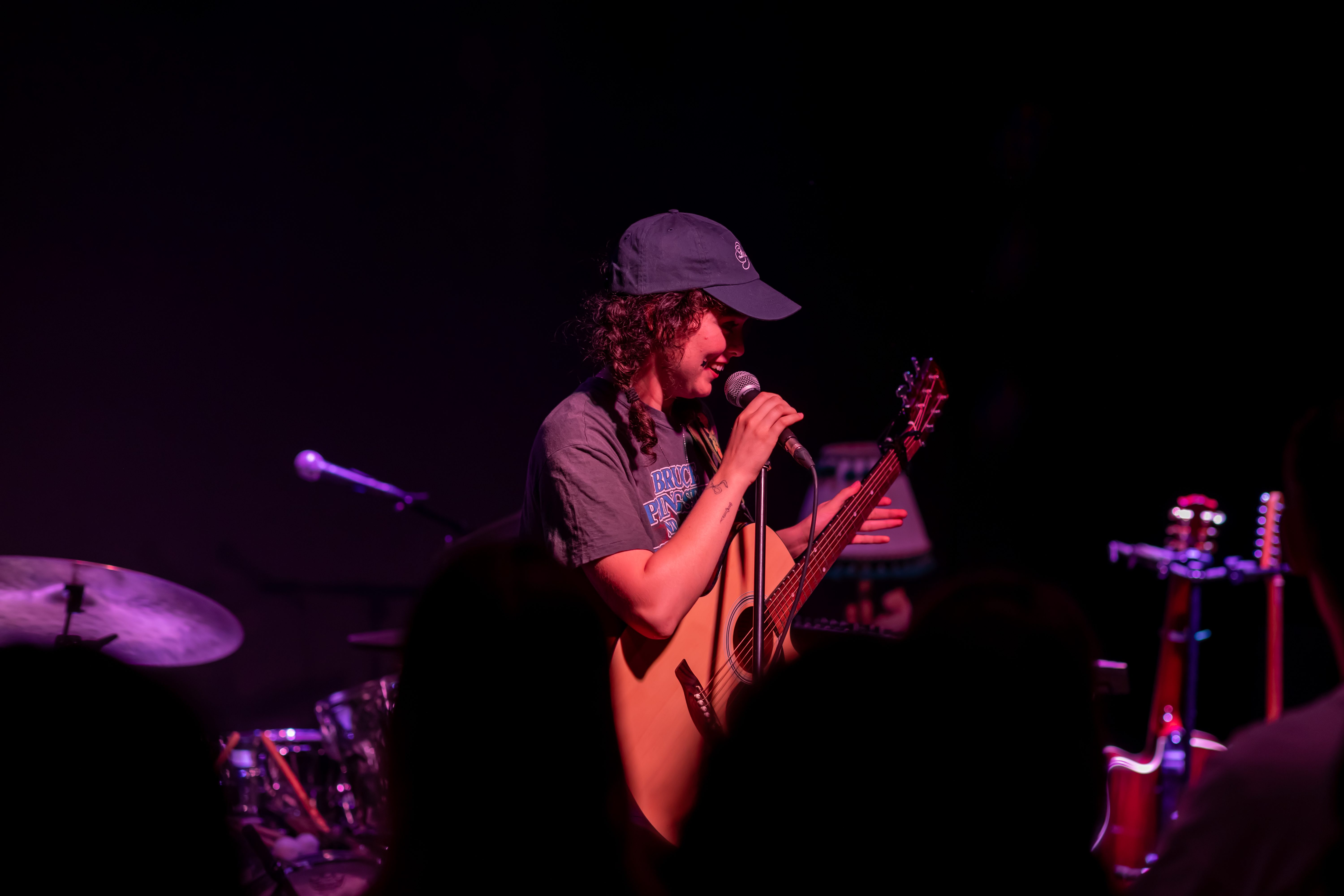 A musician performing on stage with an acoustic guitar, wearing a cap, and holding a microphone, while engaging with the audience at a live music event.