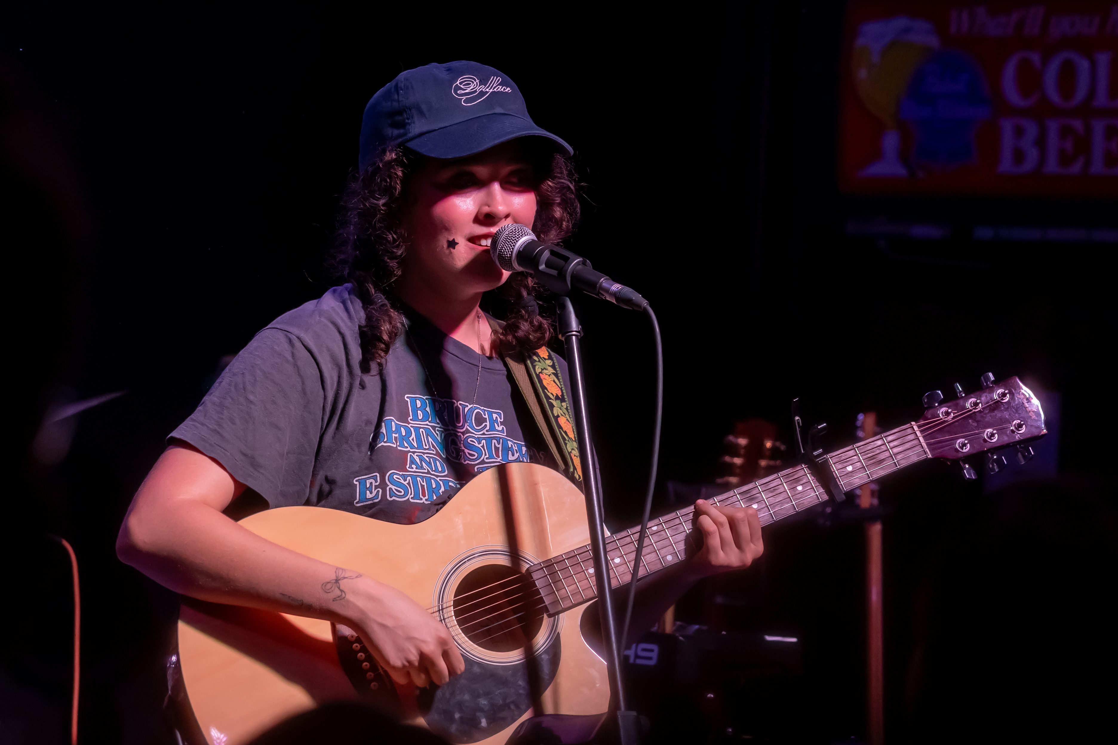 A singer performing on stage, playing an acoustic guitar and wearing a baseball cap with curly hair visible.