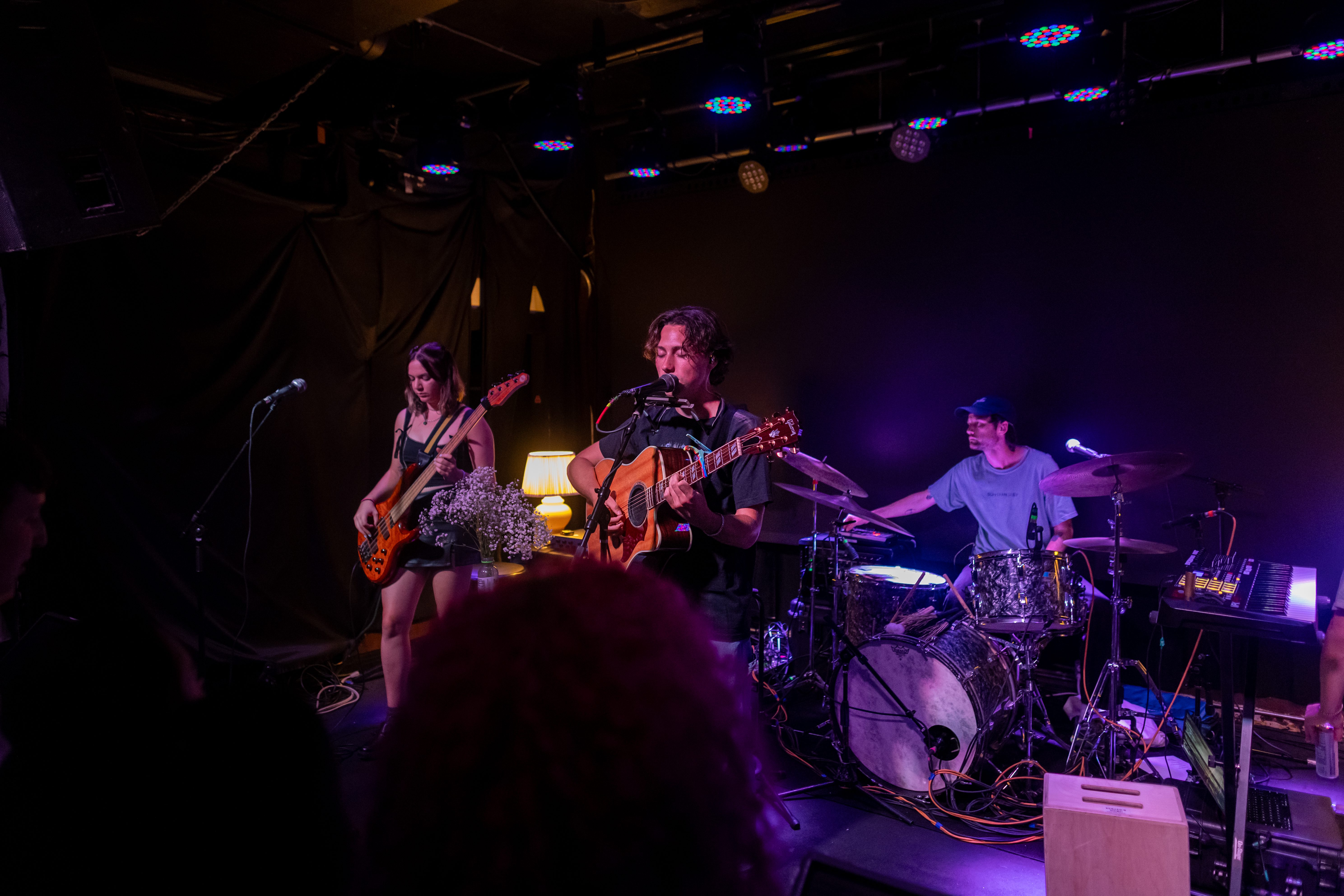 A musician playing an acoustic guitar on stage, accompanied by a bassist and a drummer, with colorful lights in the background.