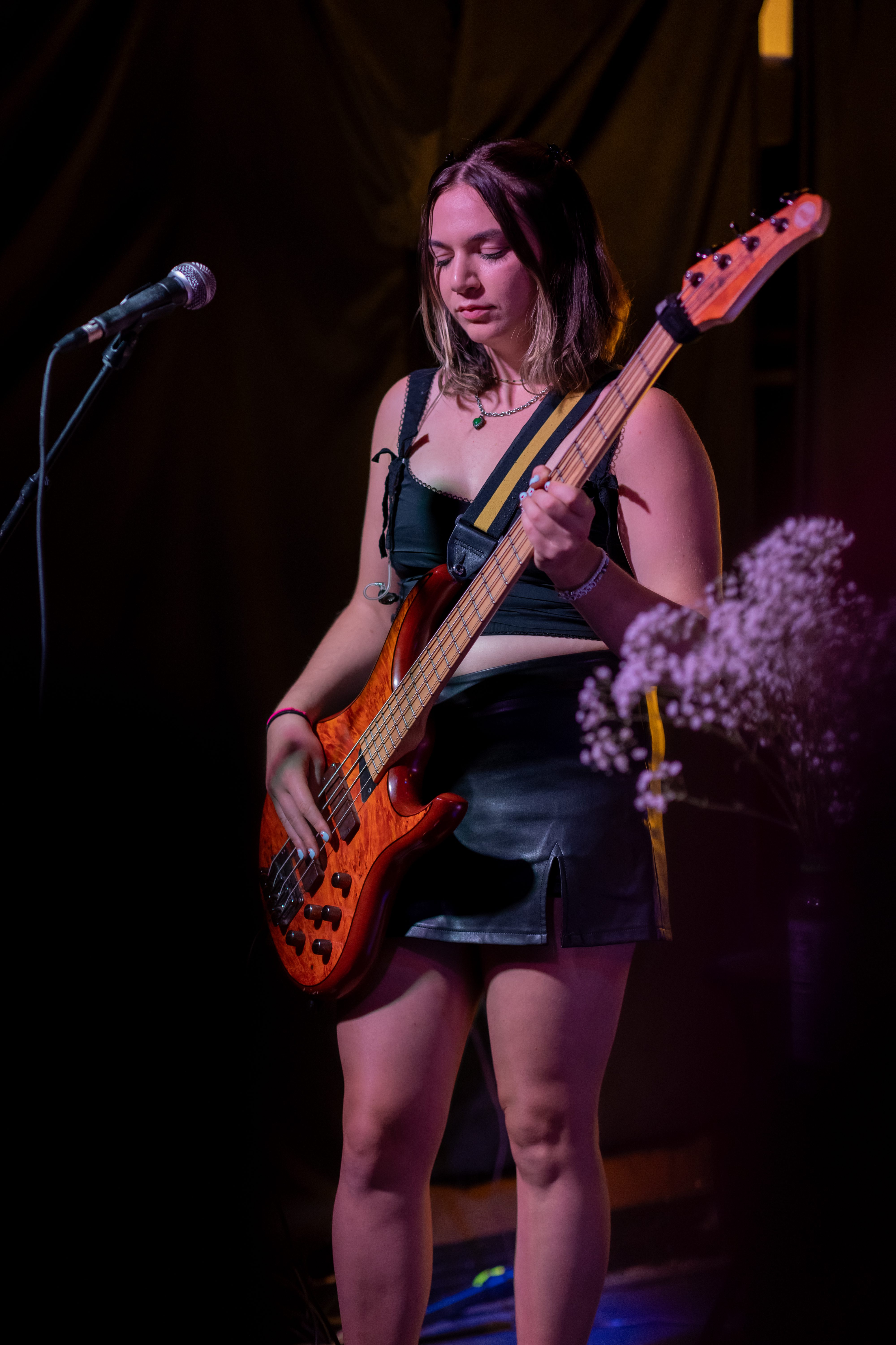 A musician playing a bass guitar on stage, wearing a black top and skirt, with a microphone in the background.