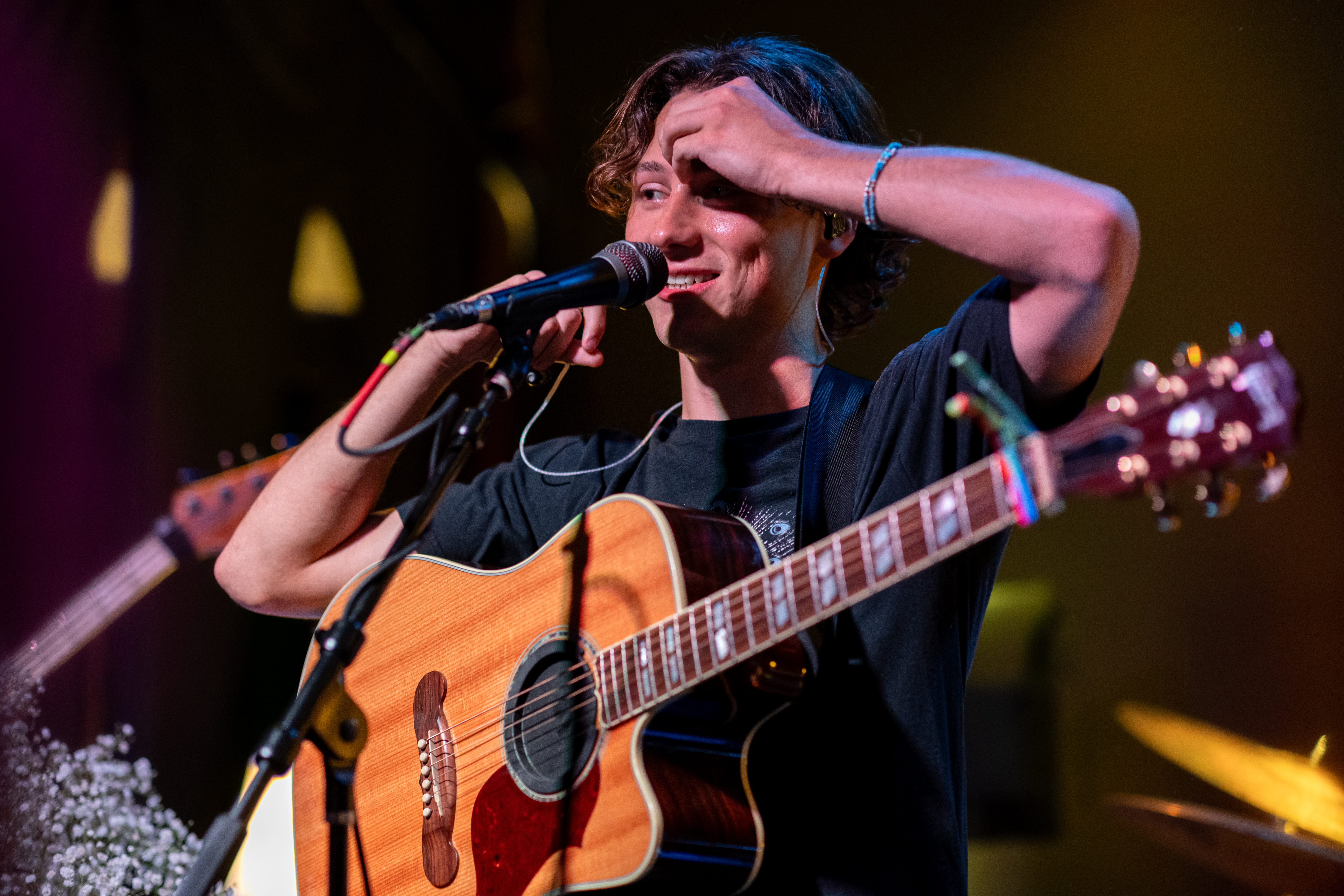 A young male musician with curly hair performs on stage, holding an acoustic guitar and smiling while speaking into a microphone.
