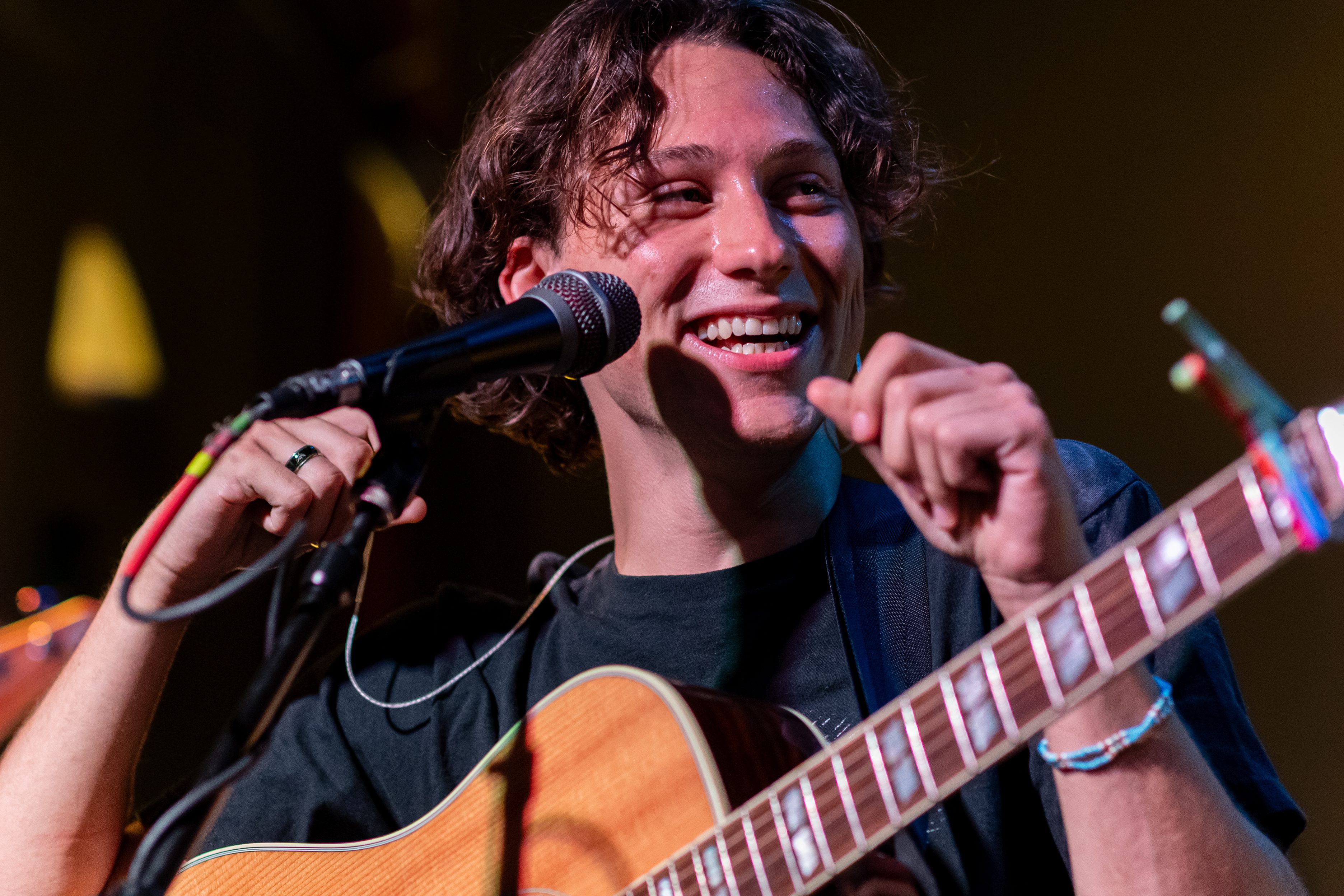 A smiling musician with curly hair playing an acoustic guitar and speaking into a microphone during a performance.