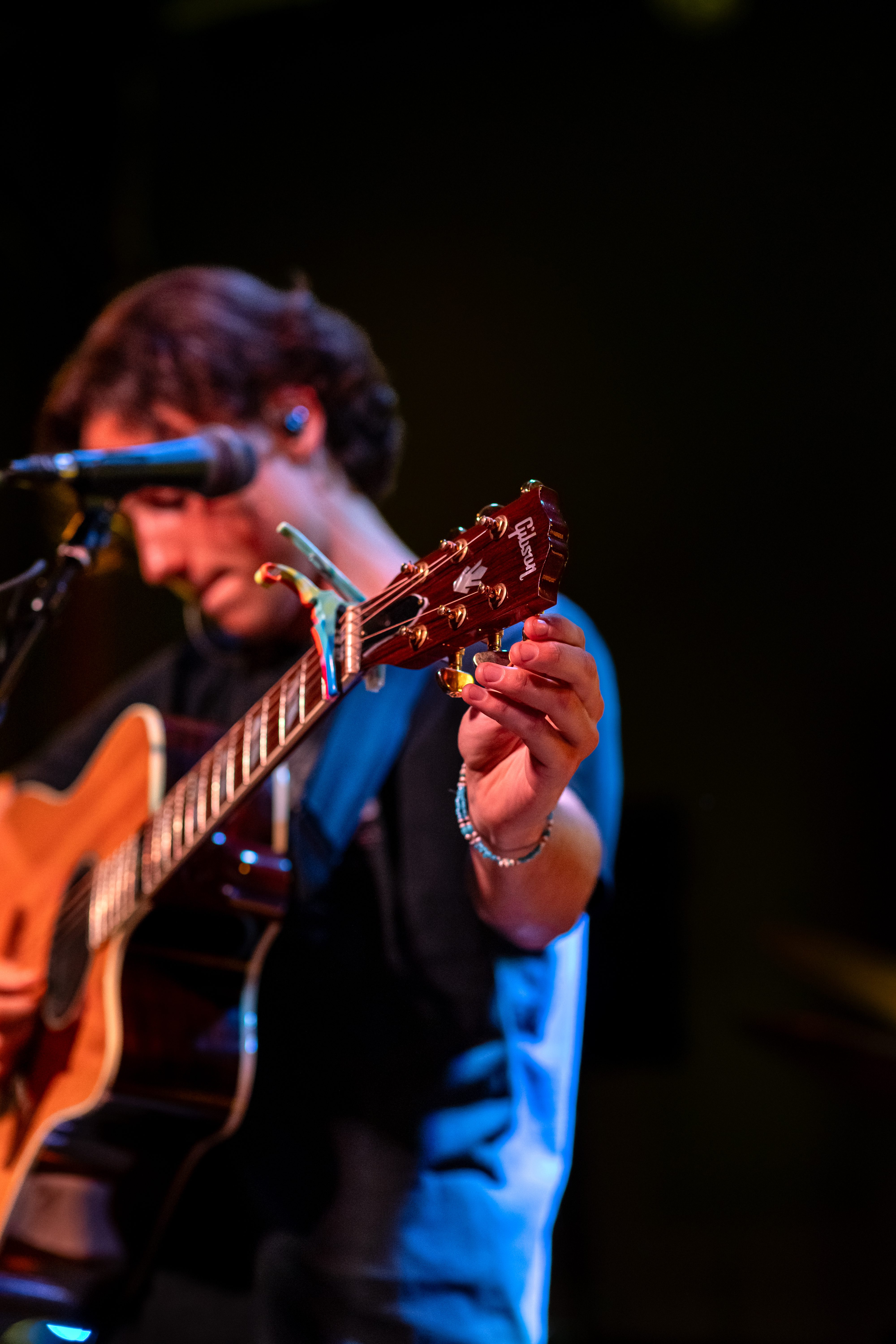 A close-up of a person's hand tuning an acoustic guitar while performing on stage, with a microphone visible.
