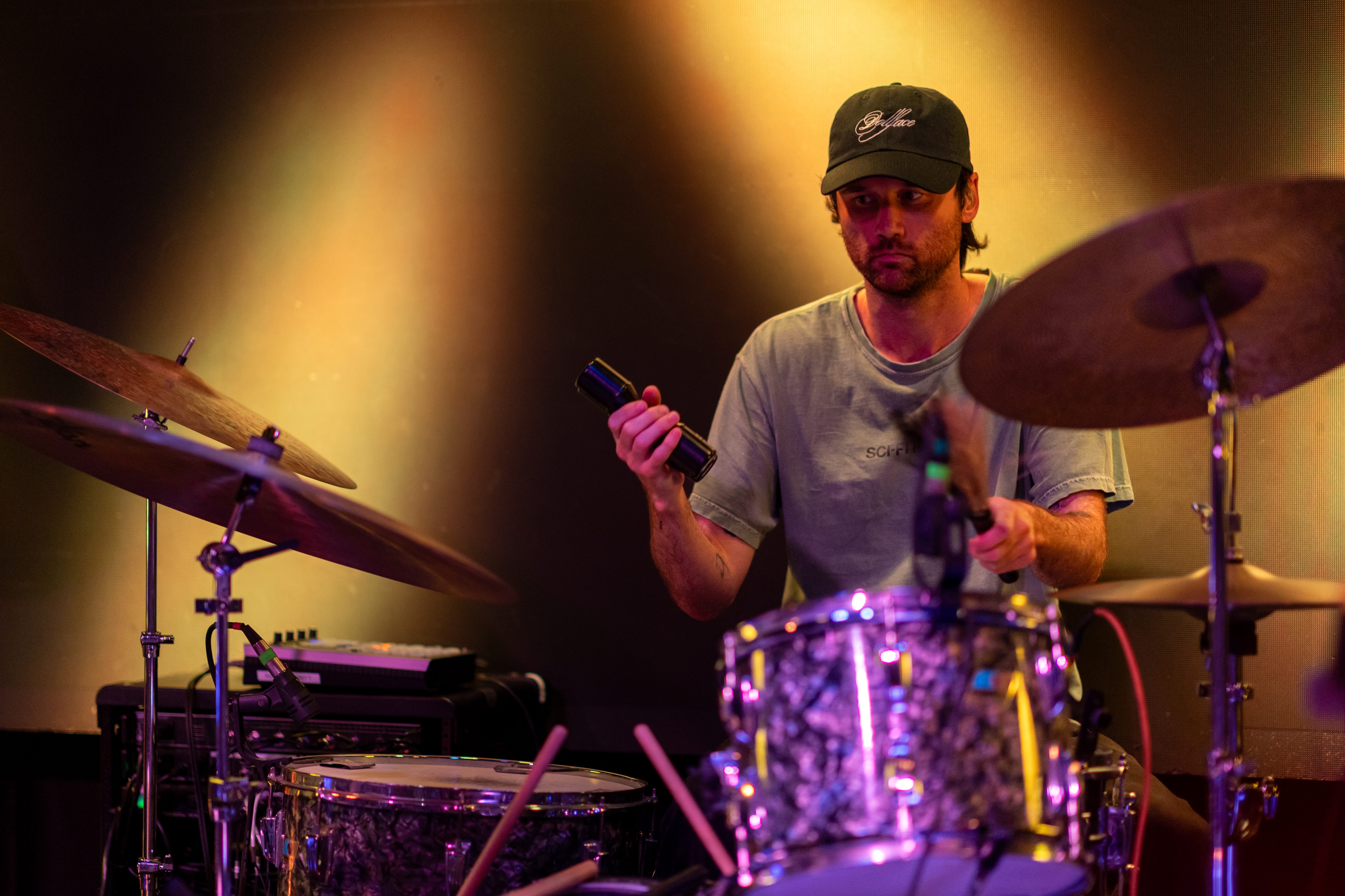 A drummer performing on stage, holding a microphone and focused on playing. The drummer is wearing a cap and a casual shirt, surrounded by drum set equipment under vibrant stage lighting.