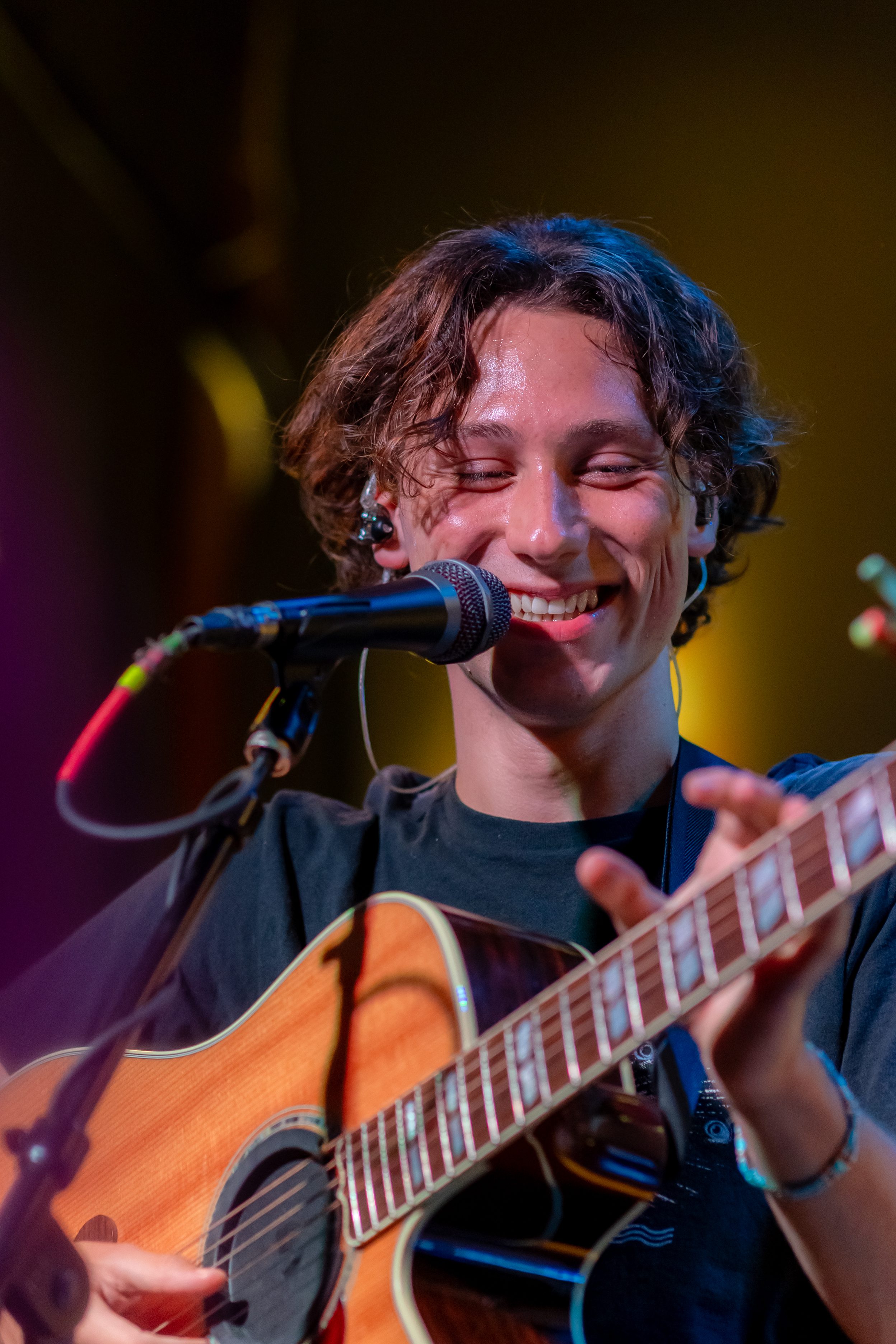 A young man with curly hair smiles while playing an acoustic guitar on stage, holding a microphone close to his mouth.
