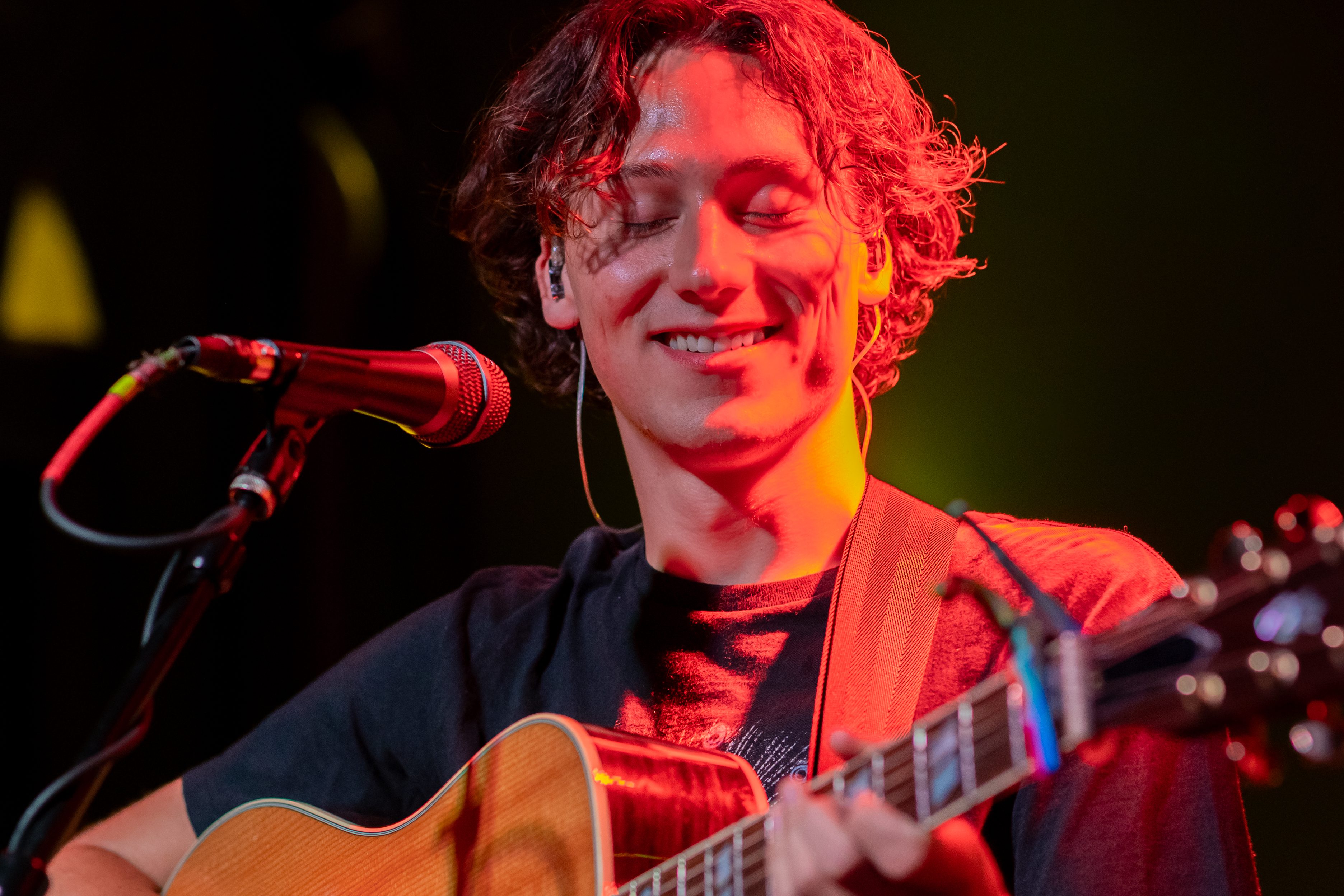 A young man with curly hair smiles while playing an acoustic guitar on stage, with a microphone in front of him and colorful stage lighting creating a vibrant atmosphere.