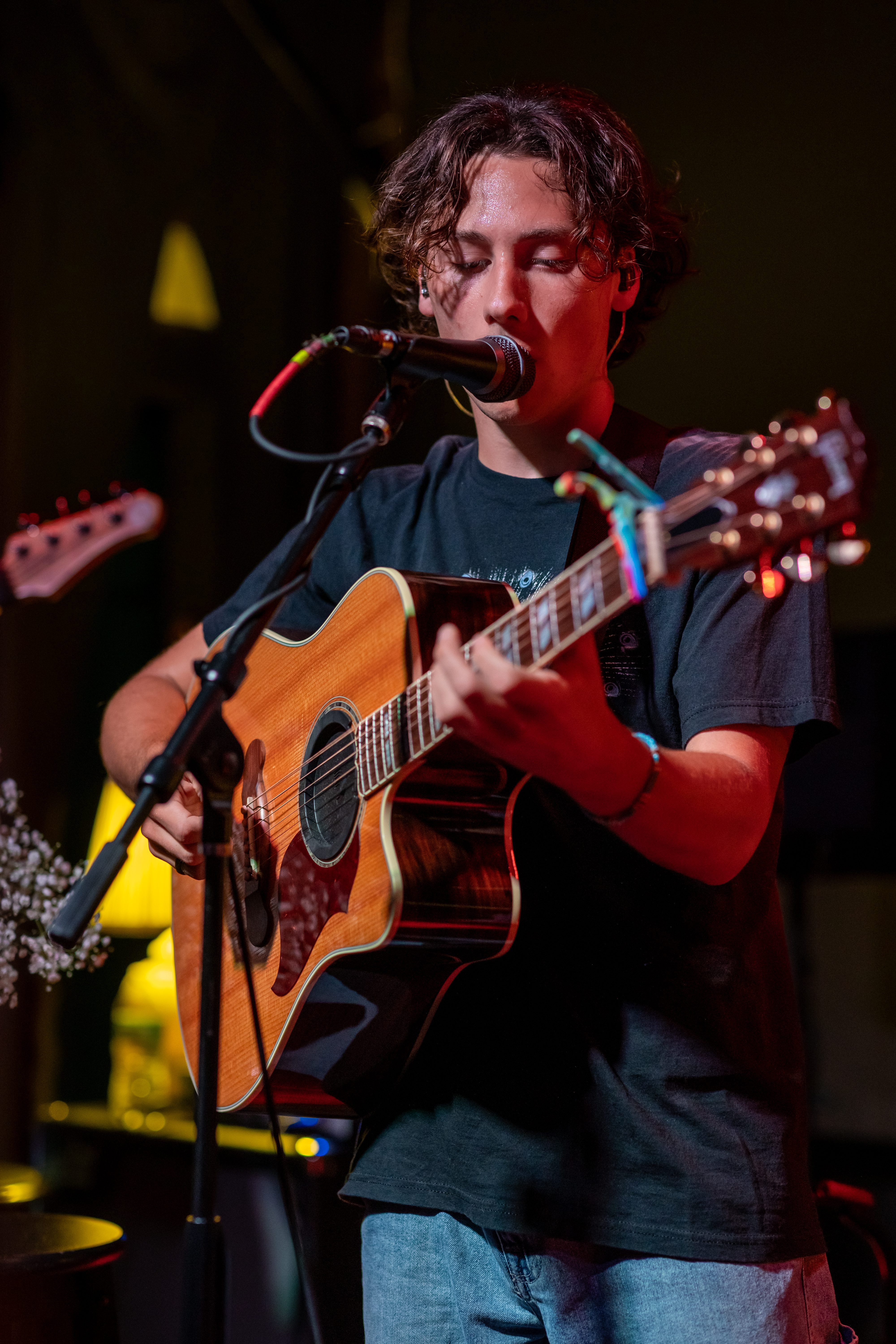 A young musician playing an acoustic guitar on stage while singing into a microphone, with colorful stage lighting and a microphone stand in front of him.