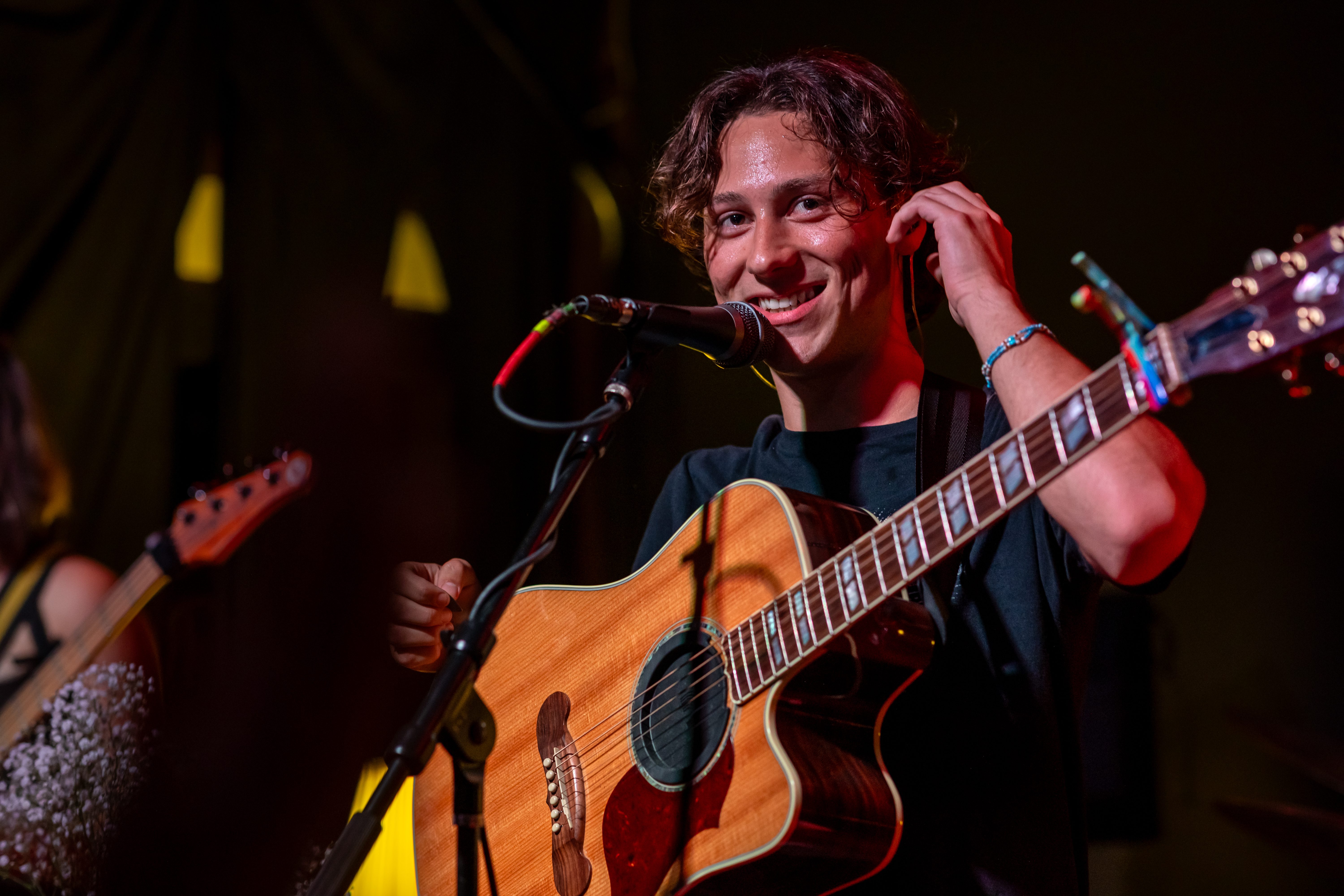 A young musician smiling while performing on stage, holding an acoustic guitar and standing in front of a microphone.