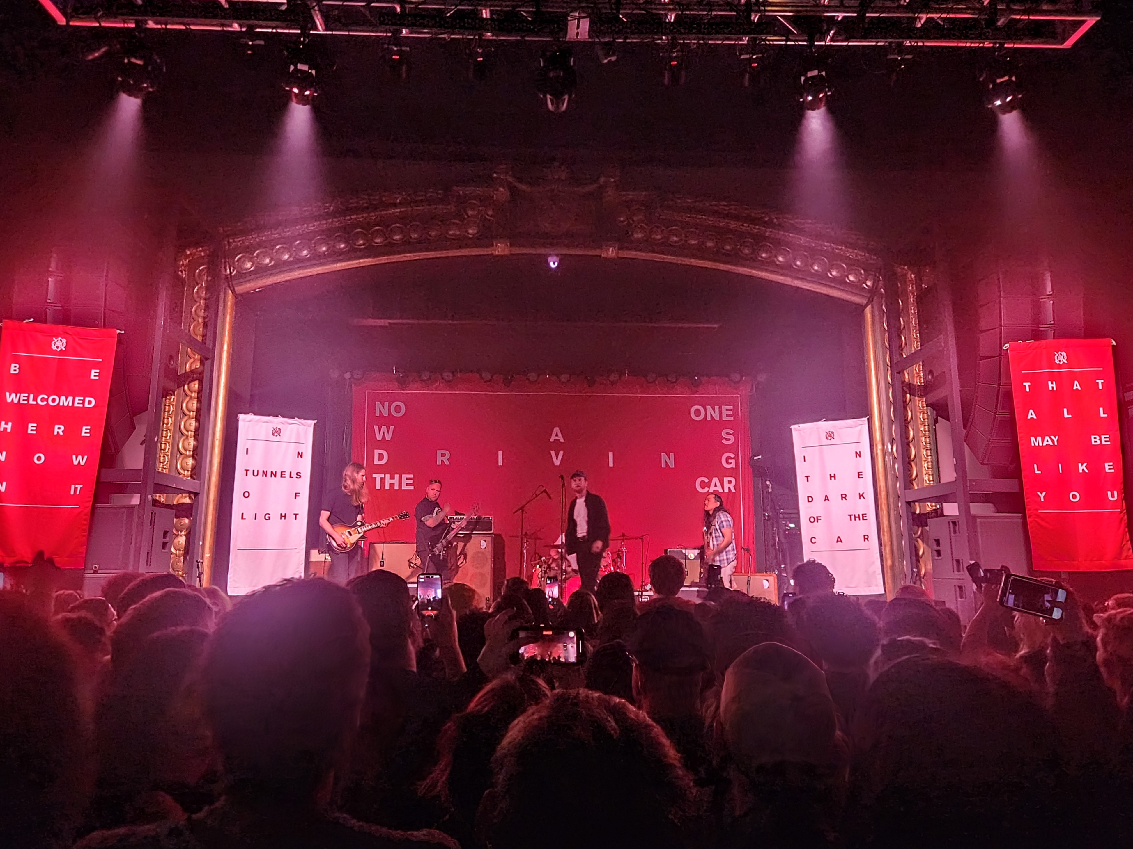 A live performance by La Dispute featuring a vibrant stage setup with banners from their album 'No One Was Driving The Car,' amidst an energetic crowd in a venue.
