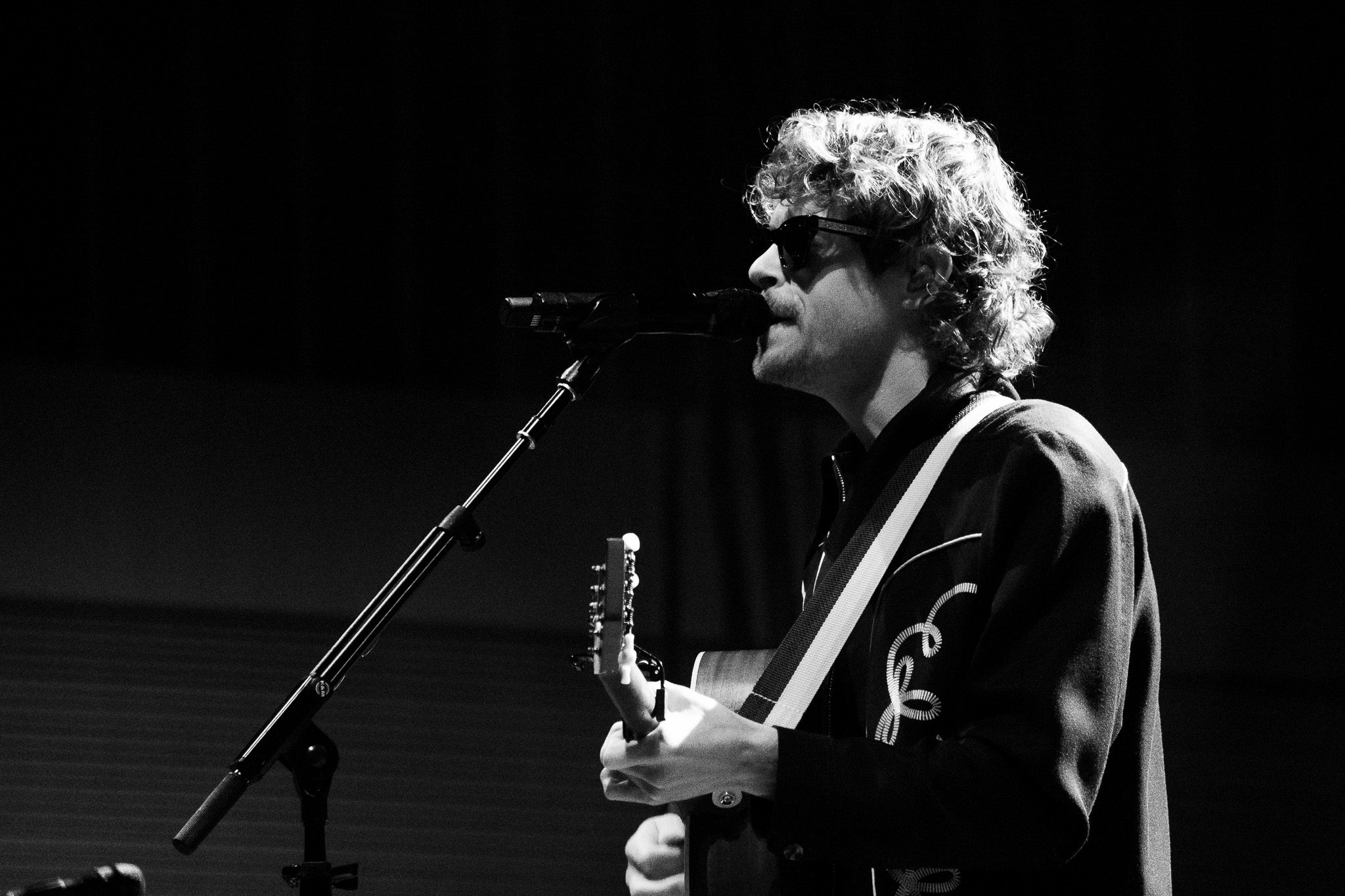 A musician performing on stage with a guitar and microphone, wearing sunglasses and a black western-style jacket, captured in black and white.