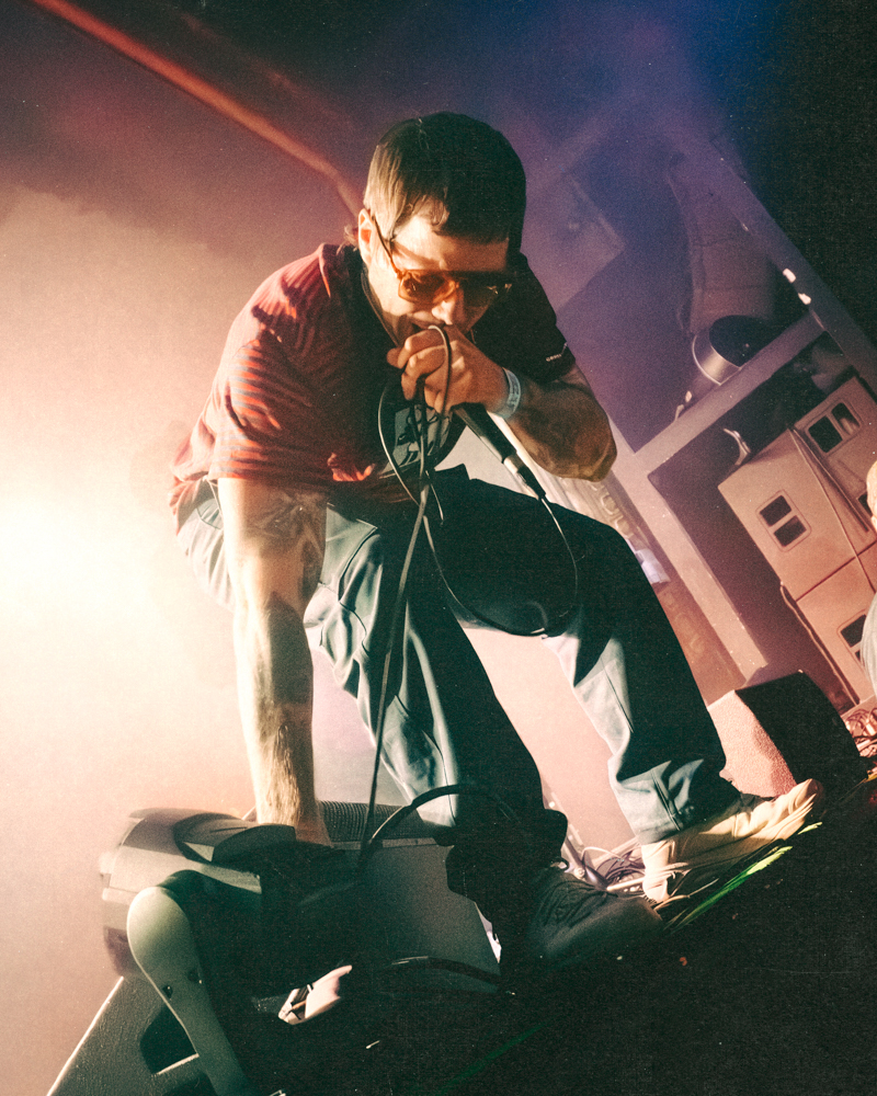 A male vocalist performing on stage with a microphone, wearing sunglasses and a striped red shirt, leaning forward with one foot on a speaker.