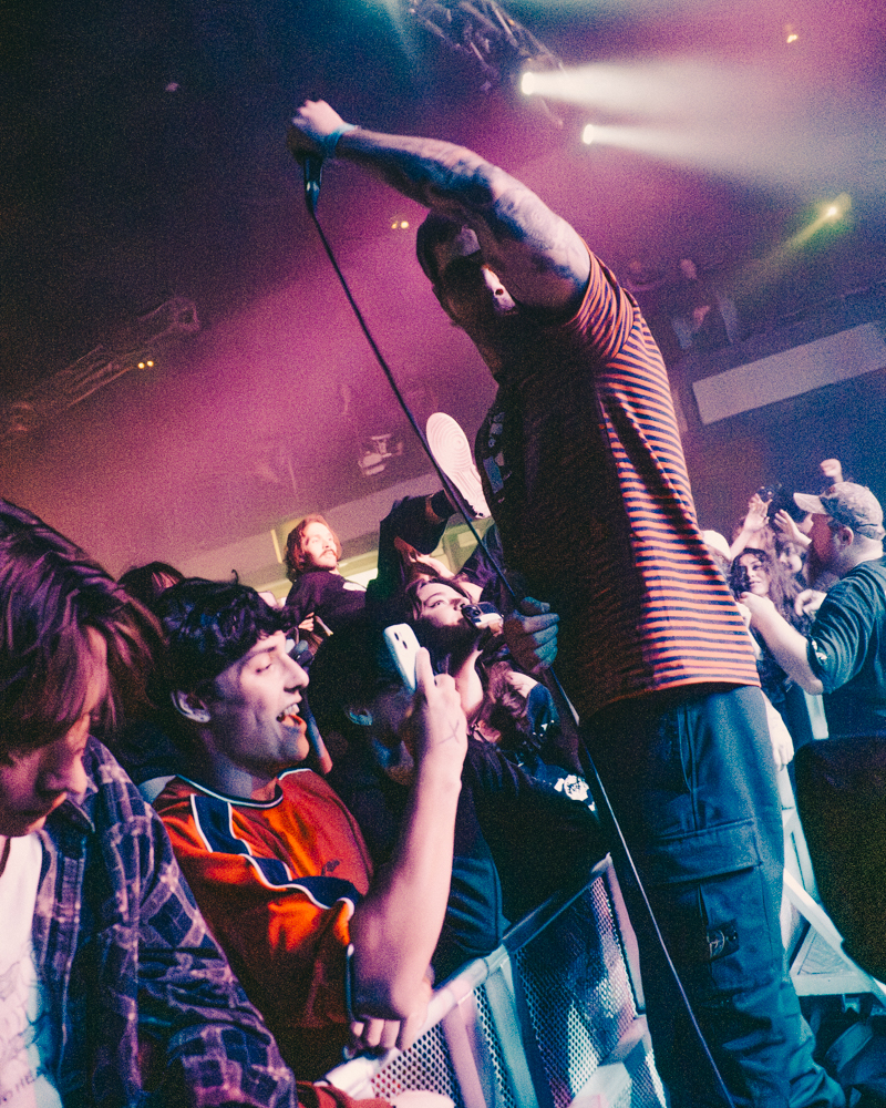 A live concert scene featuring a passionate vocalist engaging with an enthusiastic crowd, with audience members close to the stage, capturing the moment on their phones under colorful stage lighting.