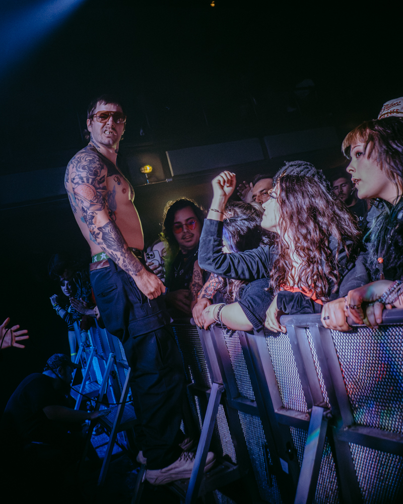 A shirtless performer with tattoos engages with excited fans at a concert, standing on a barricade while fans reach out and cheer.