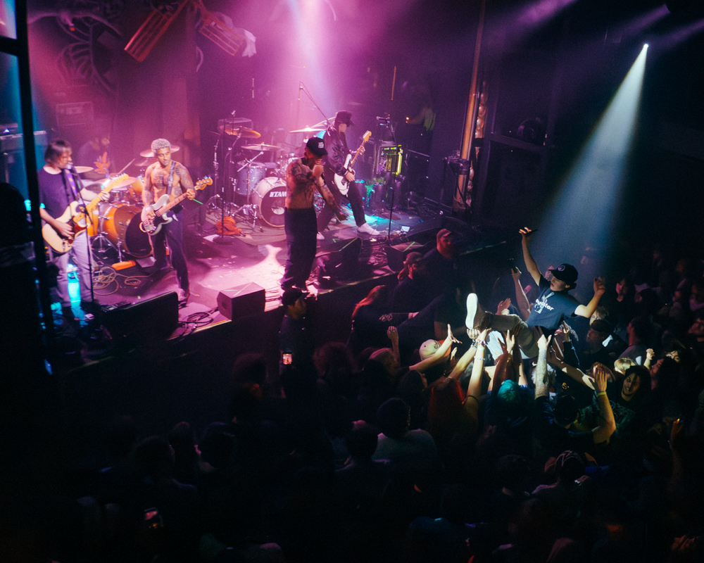 A live concert scene featuring a band performing on stage with colorful lighting, while enthusiastic crowd members are crowd-surfing and enjoying the energetic atmosphere.