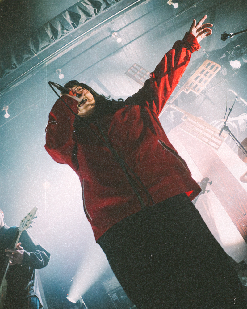 A singer performing on stage, wearing a red jacket, with dramatic stage lighting creating an energetic atmosphere.